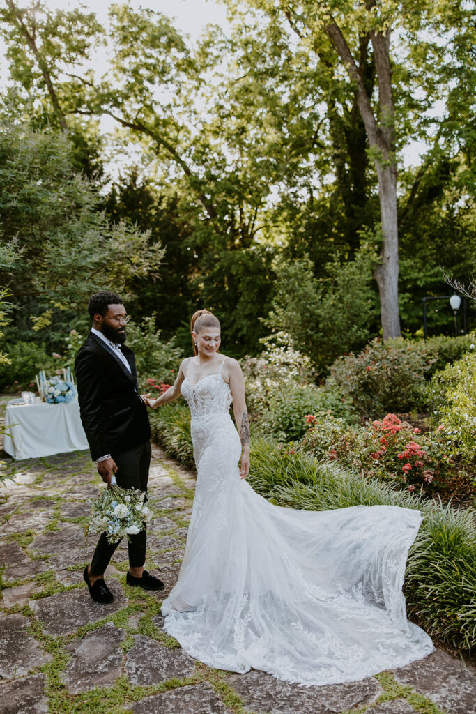 A couple in wedding attire stands on a stone path in a lush garden, holding hands and looking at each other.