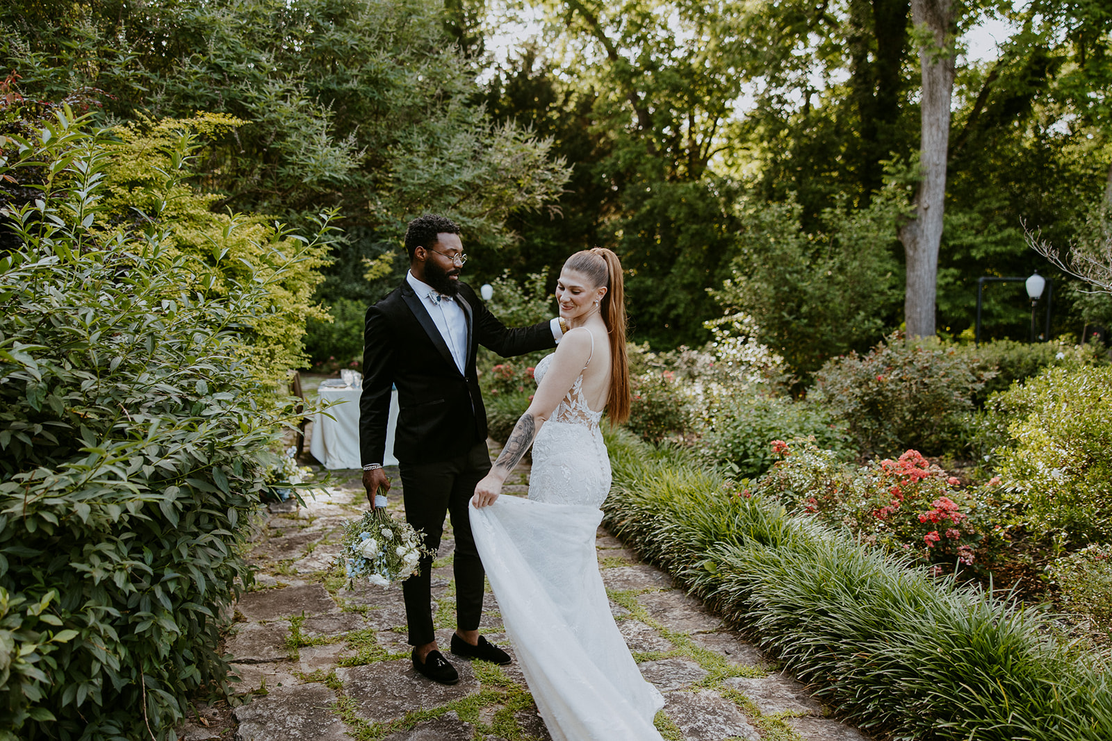 A couple in wedding attire stands on a stone path in a lush garden, holding hands and looking at each other.