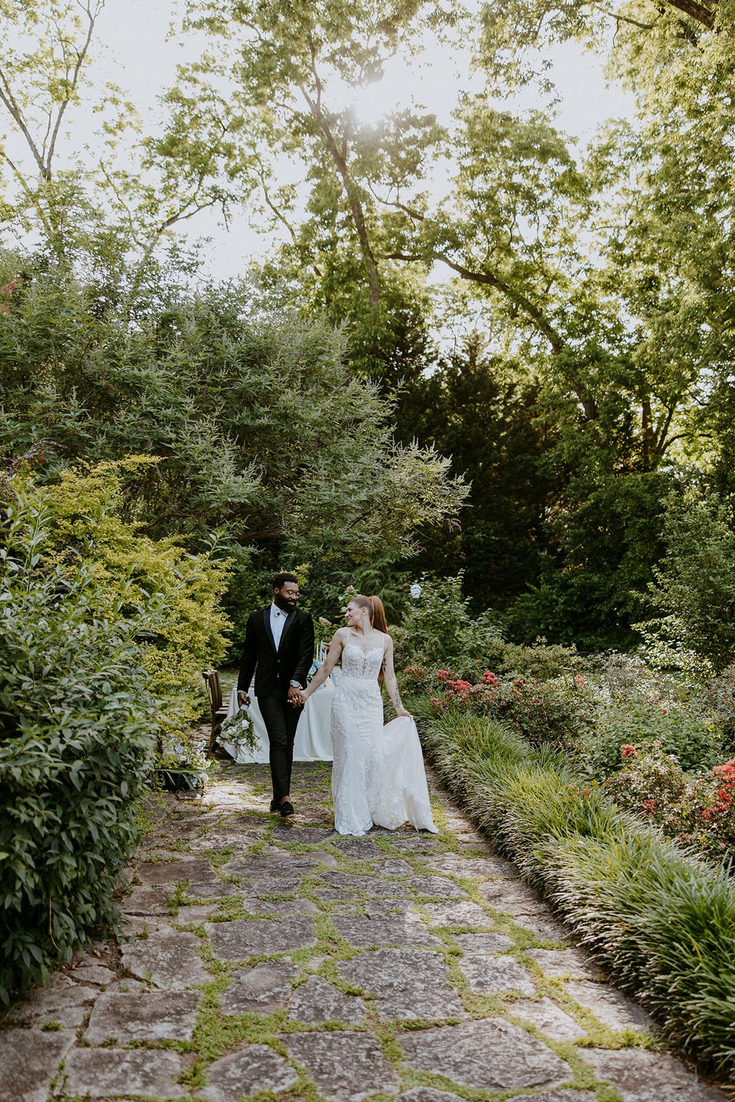 A couple in wedding attire stands on a stone path in a lush garden, holding hands and looking at each other.