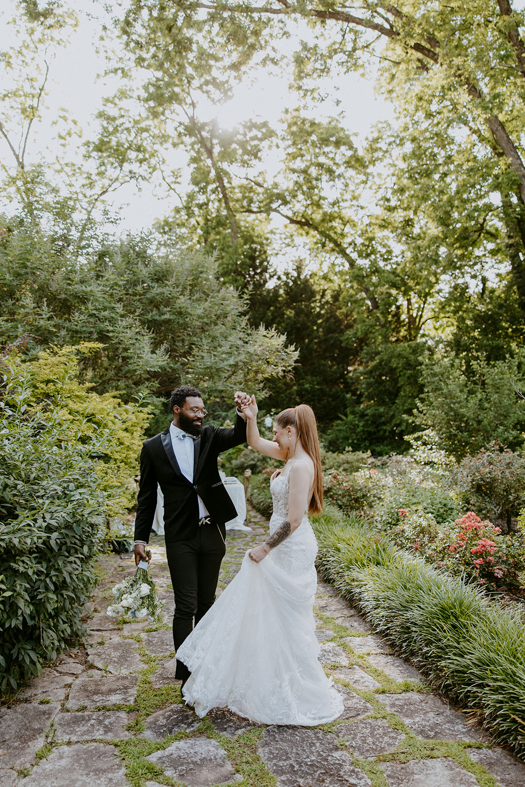 A couple in wedding attire stands on a stone path in a lush garden, holding hands and looking at each other.