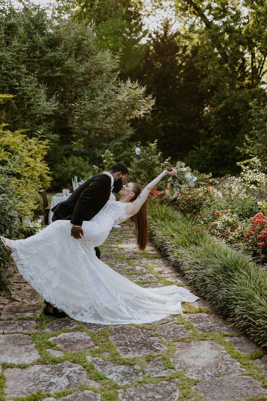 A couple in wedding attire stands on a stone path in a lush garden, holding hands and looking at each other.