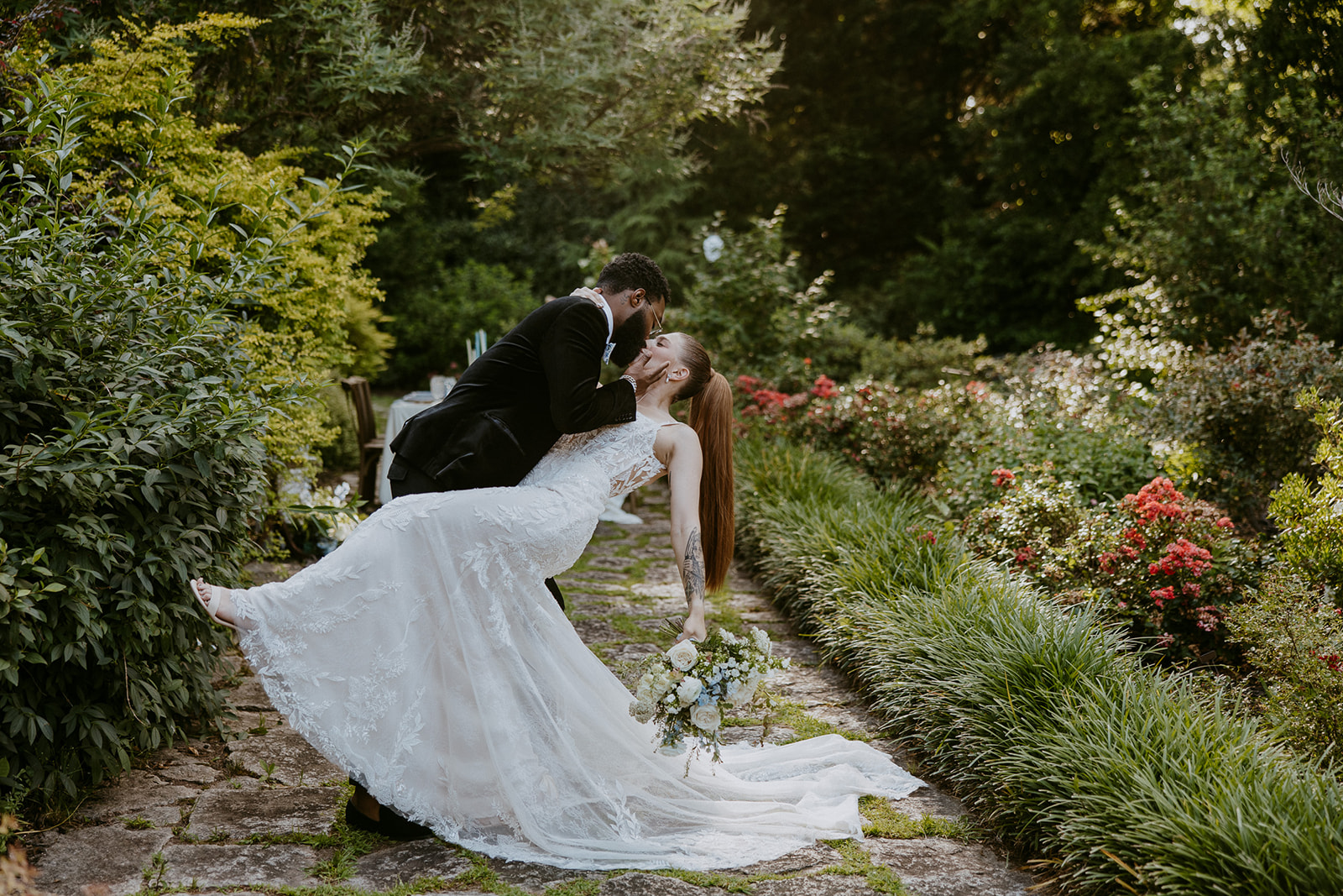 A couple in wedding attire stands on a stone path in a lush garden, holding hands and looking at each other.