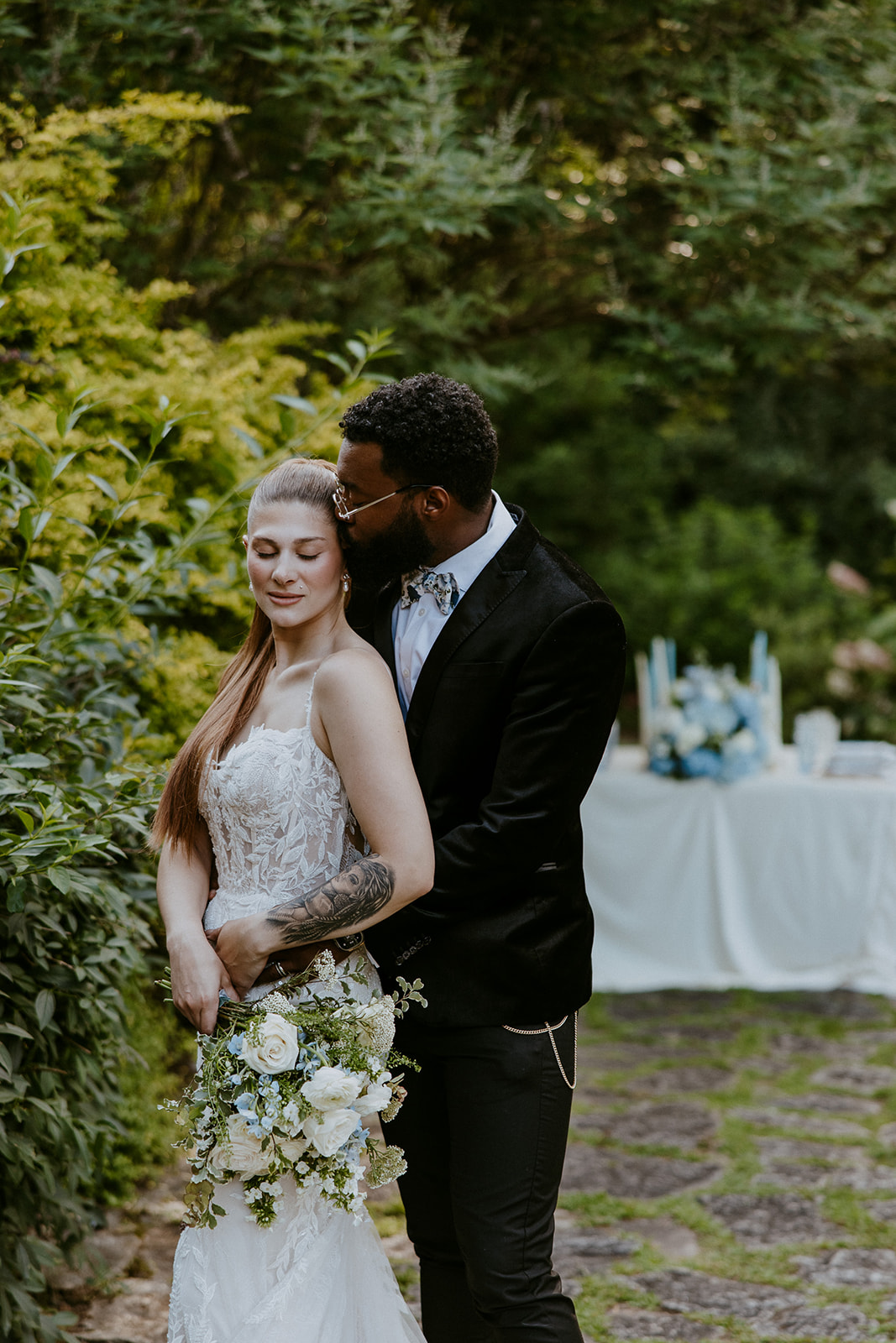 A couple stands outdoors in a garden setting, at Wildflower 301 wearing wedding attire. They are smiling and holding champagne glasses. A decorated table with a blue and white cake is beside them