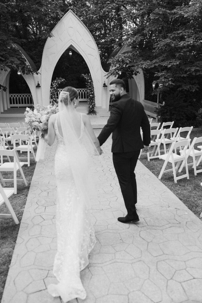 A bride and groom walk down an outdoor aisle holding hands, with empty chairs and an arch structure in the background at wildflower 301