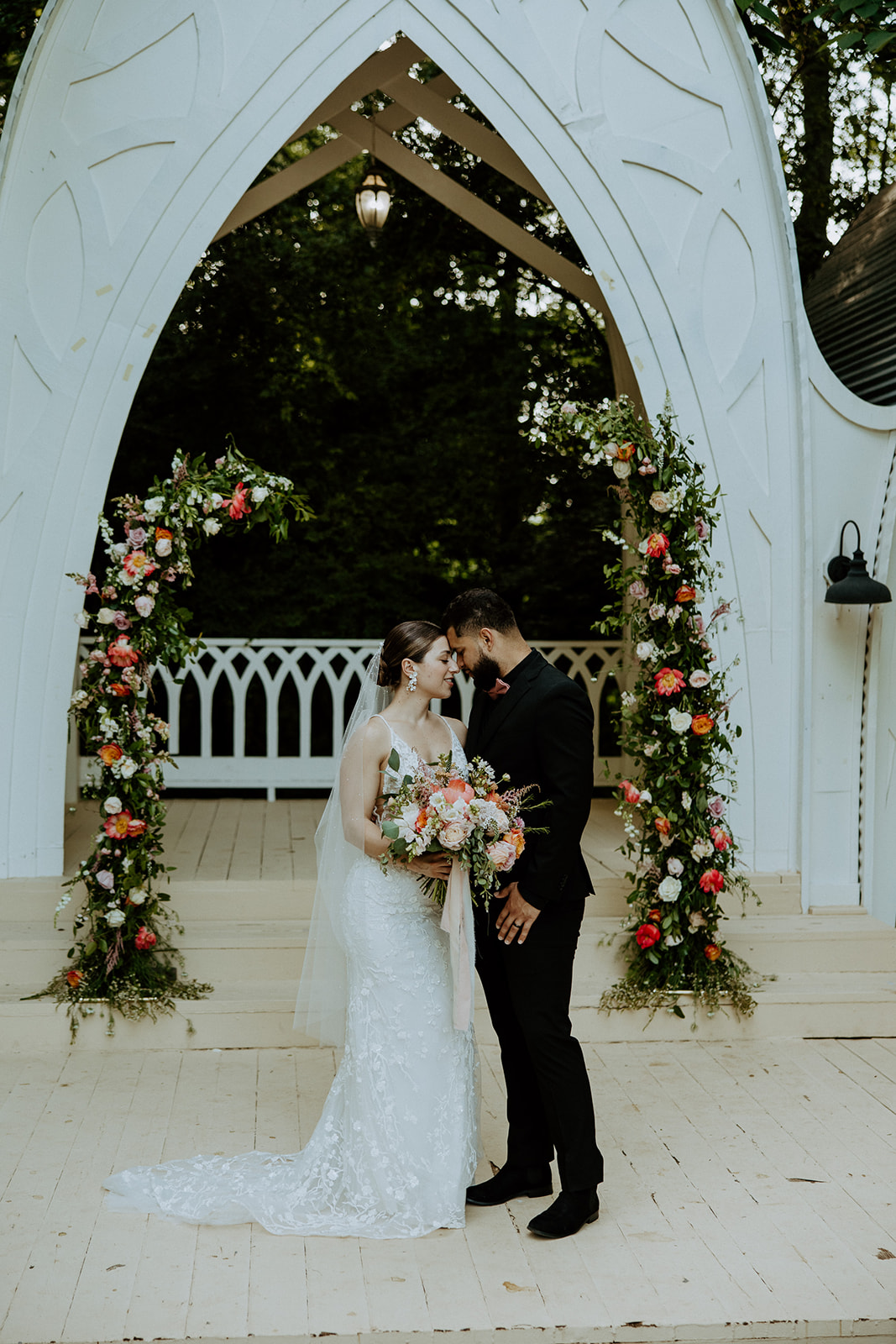 A couple in wedding attire share a kiss. The bride wears a veil and earrings, while the groom is in a dark suit. Flowers and greenery are visible in the background.