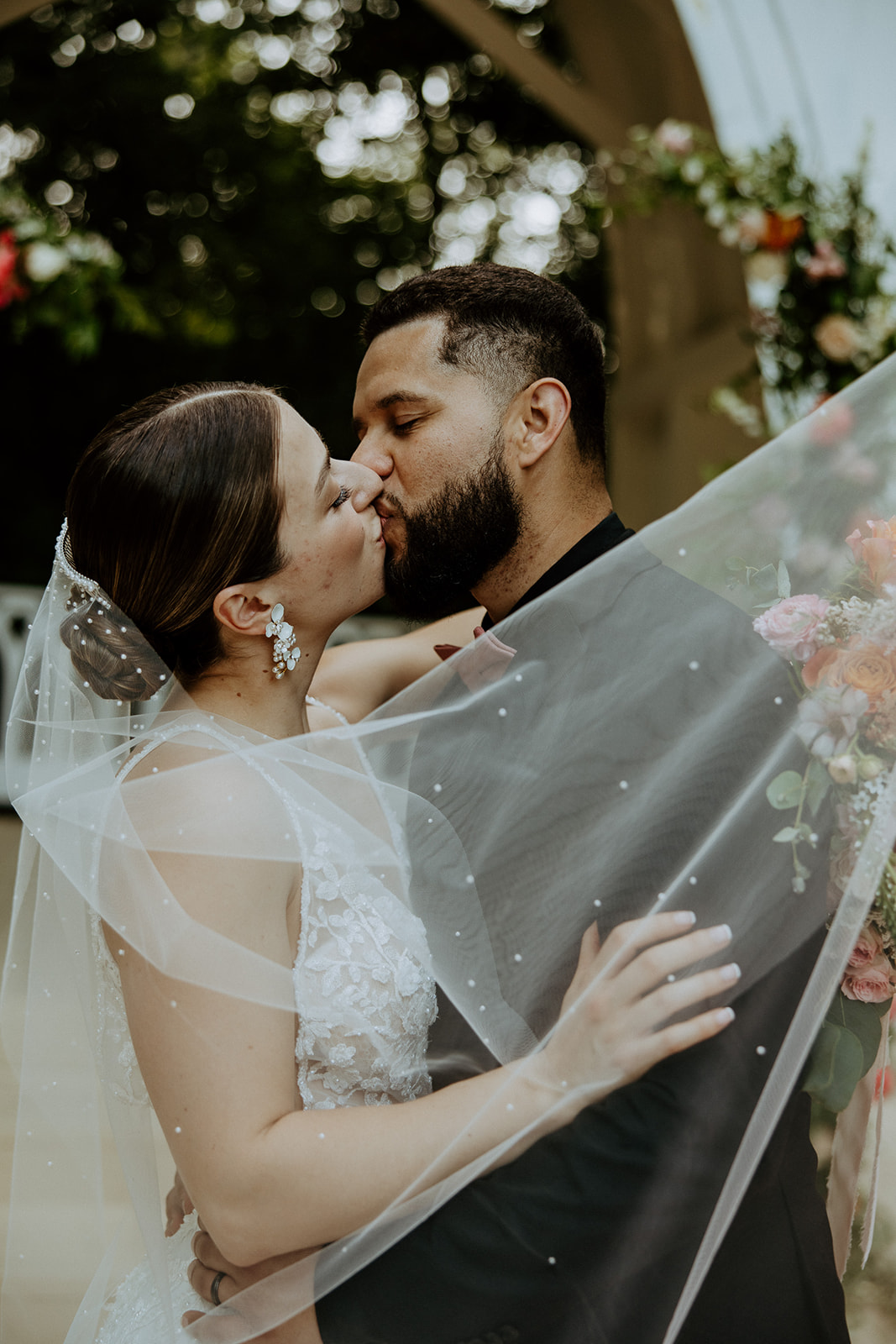 A bride and groom share a kiss under a sheer veil, with floral decorations in the background.