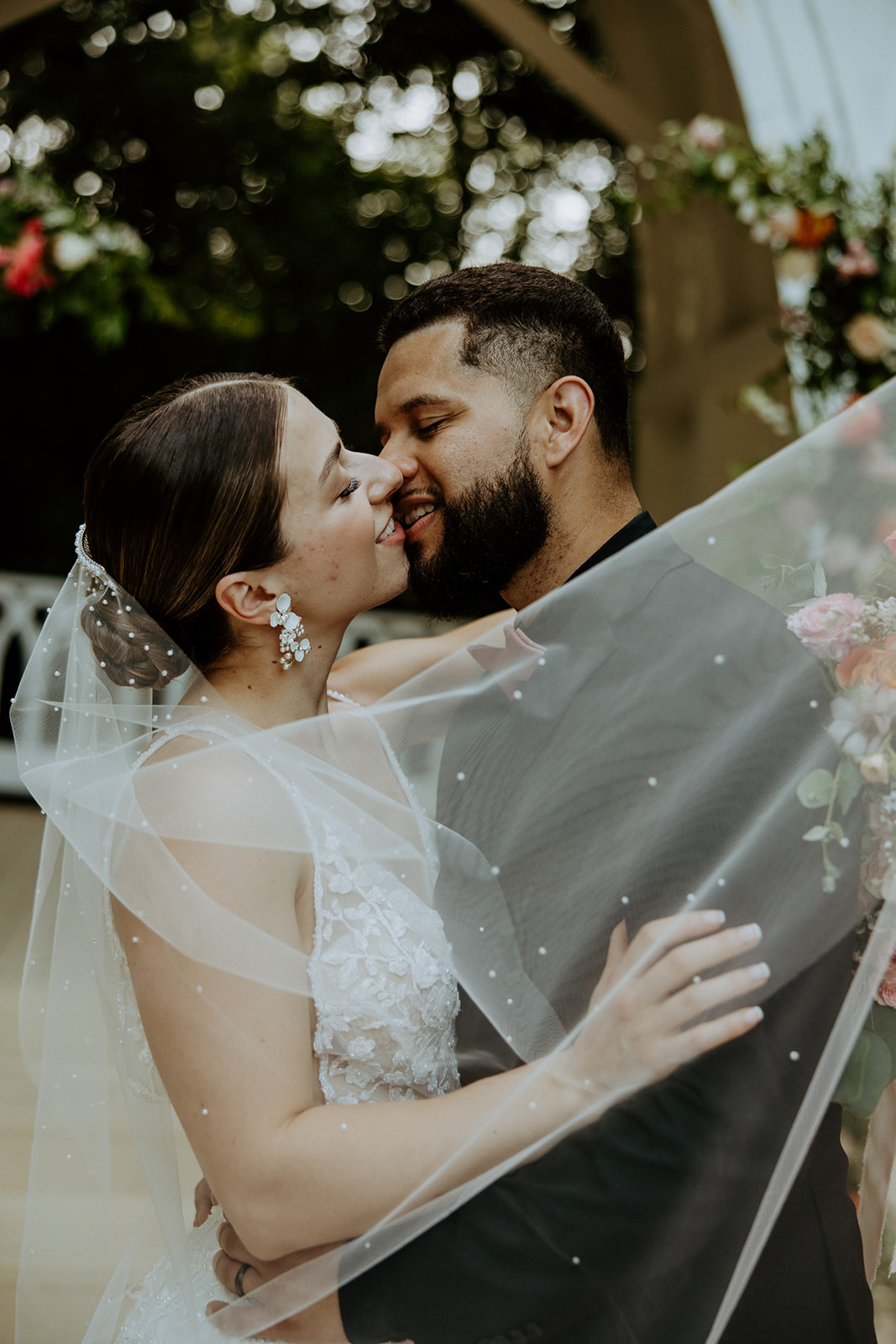 A couple in wedding attire share a kiss. The bride wears a veil and earrings, while the groom is in a dark suit. Flowers and greenery are visible in the background.