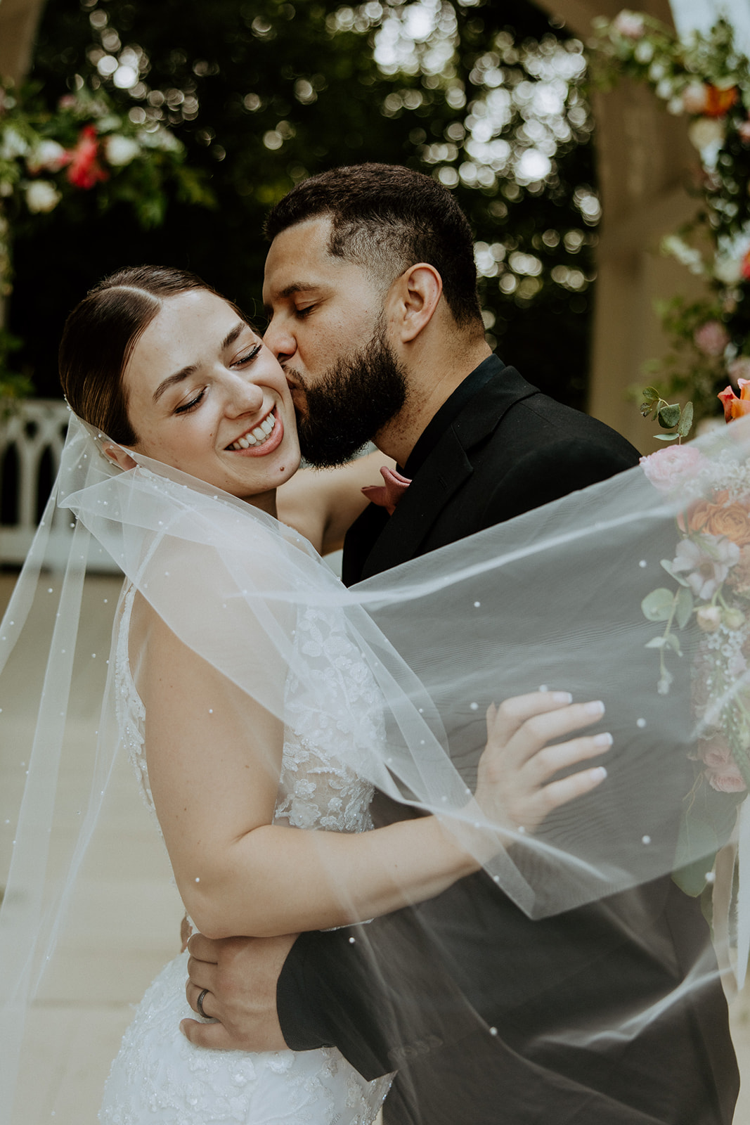 A bride and groom share a kiss under a sheer veil, with floral decorations in the background.