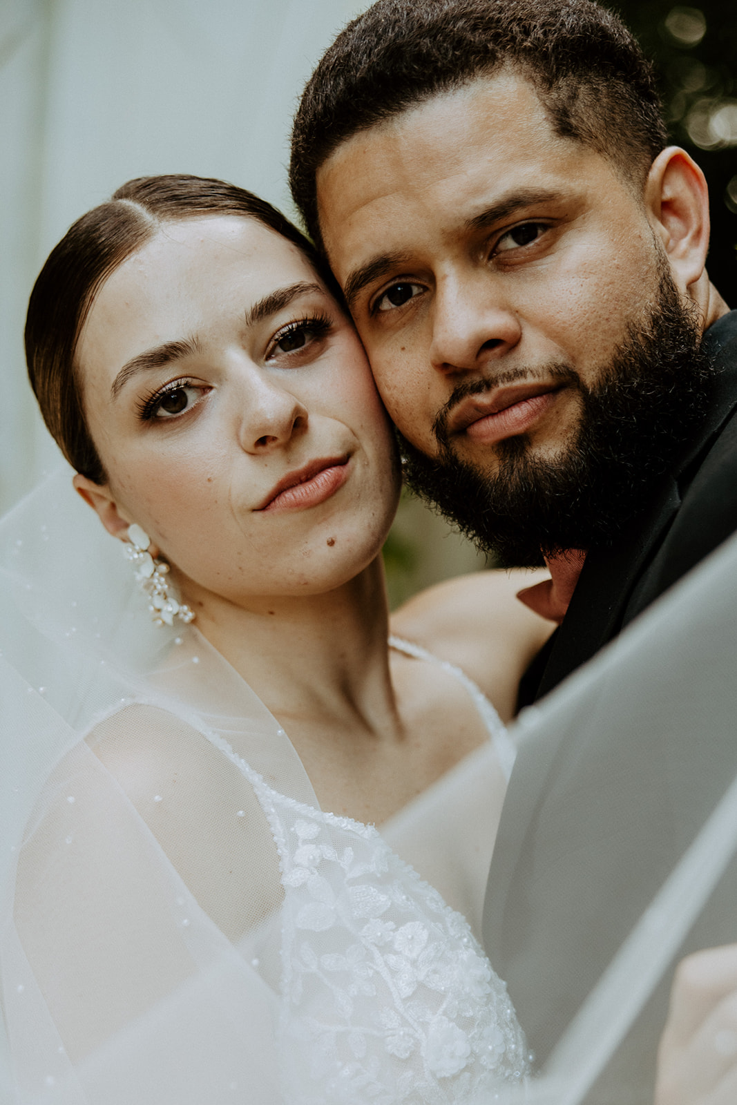 A couple in wedding attire share a kiss. The bride wears a veil and earrings, while the groom is in a dark suit. Flowers and greenery are visible in the background.