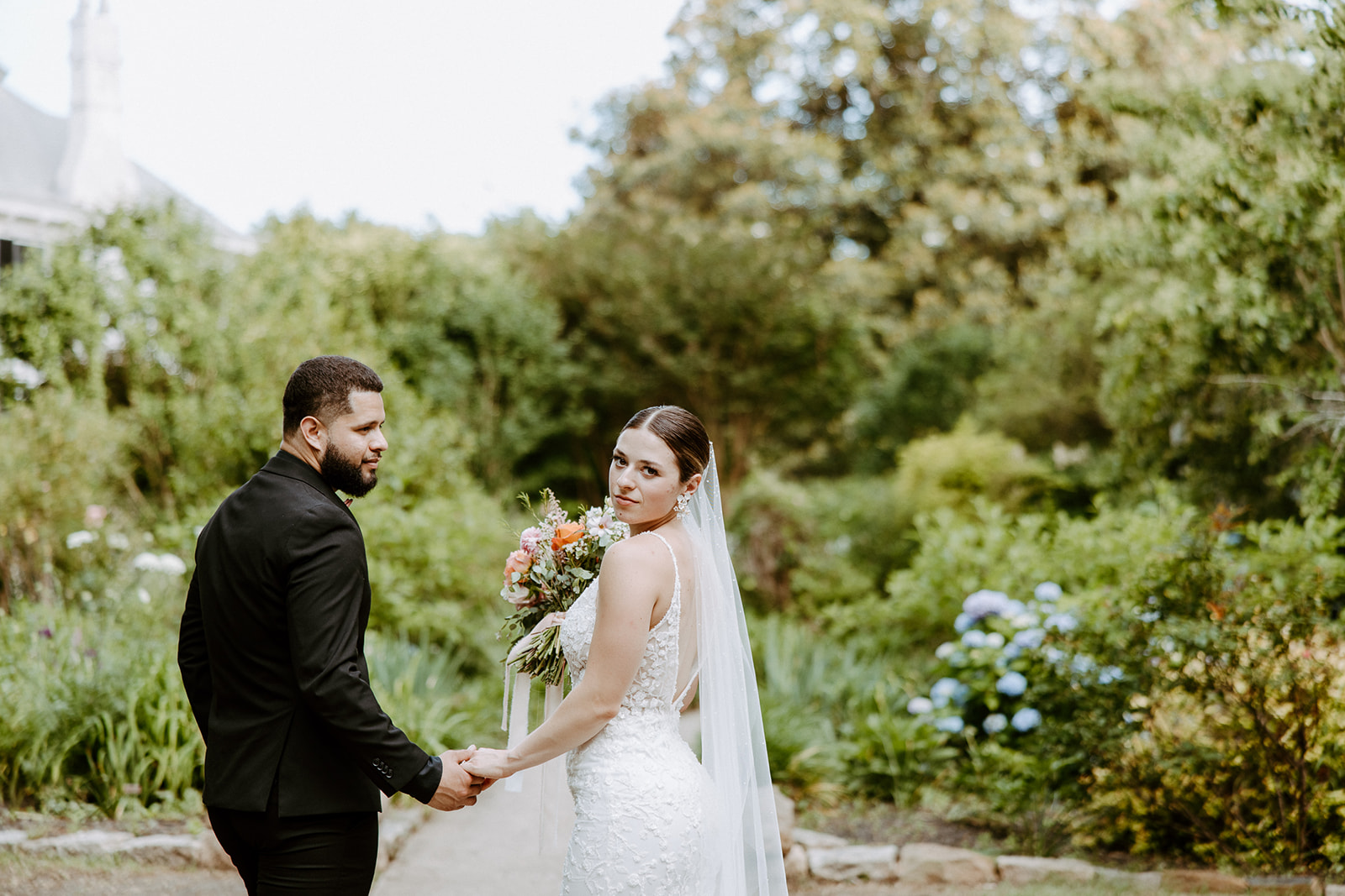 A bride in a white dress holding a bouquet and a groom in a black suit walk along a garden pathway at Wildflower 301