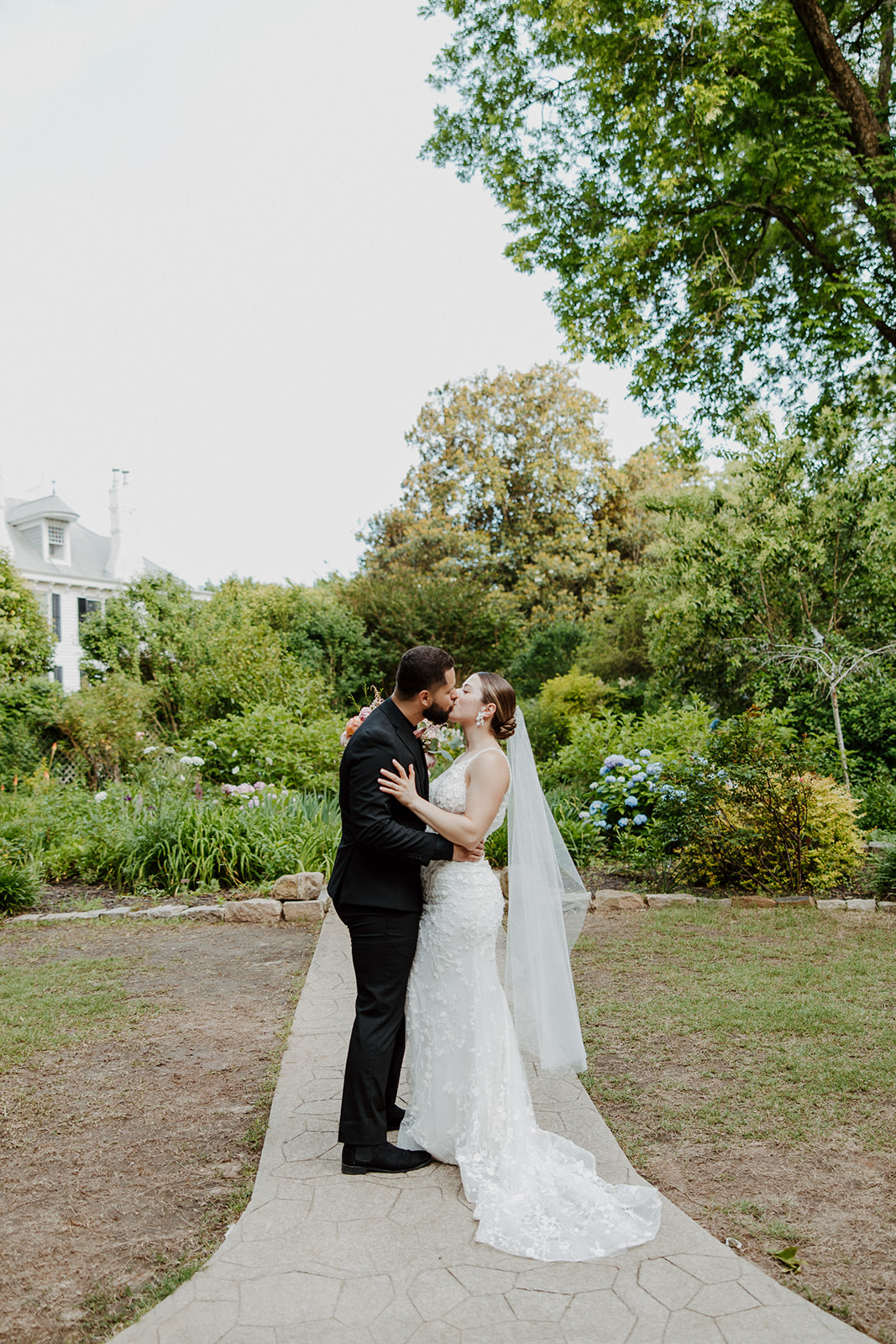 A bride in a white dress holding a bouquet and a groom in a black suit walk along a garden pathway at Wildflower 301