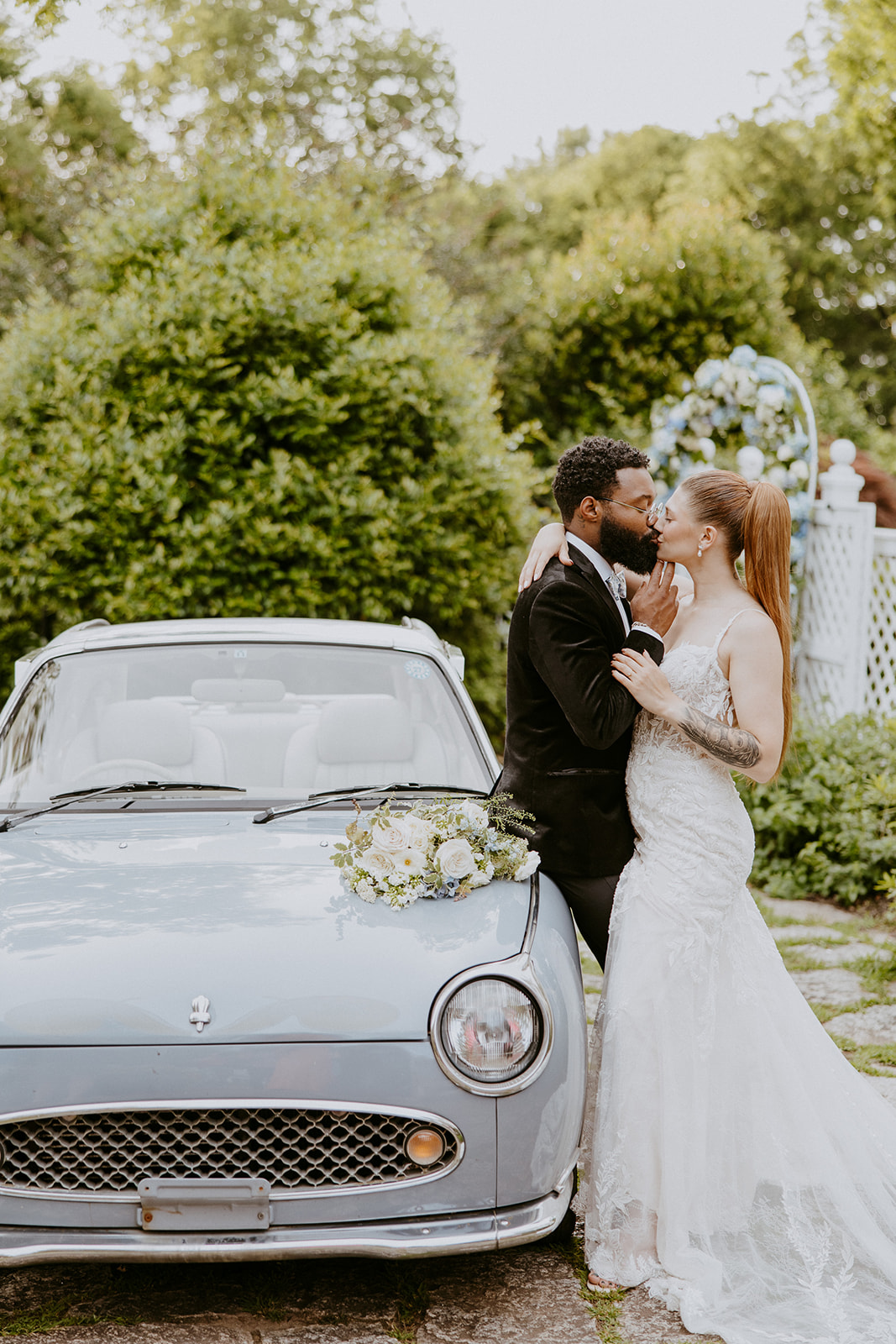 A couple stands beside a light blue vintage car. The woman in a white dress embraces the man in a black suit who holds her face gently. The car has a small bouquet on its hood. Greenery is in the background at Wildflower 301