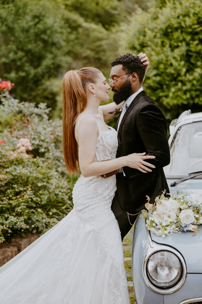 A bride in a white dress and a groom in a black suit stand close together, embracing in front of a blue vintage car with a flower arrangement on its hood. They are surrounded by greenery and flowers at Wildflower 301