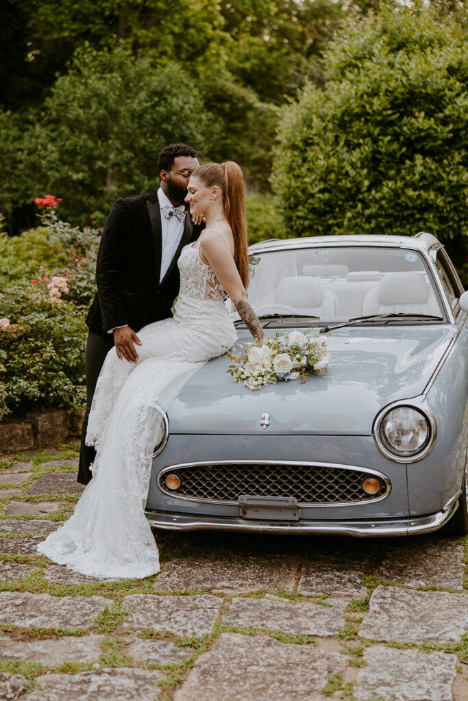 A bride in a white dress and a groom in a black suit stand close together, embracing in front of a blue vintage car with a flower arrangement on its hood. They are surrounded by greenery and flowers at Wildflower 301