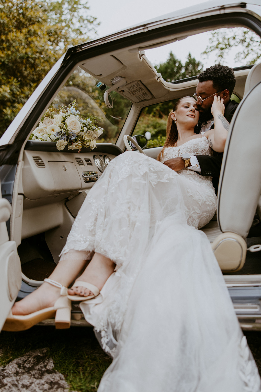 A couple stands beside a light blue vintage car. The woman in a white dress embraces the man in a black suit who holds her face gently. The car has a small bouquet on its hood. Greenery is in the background at Wildflower 301