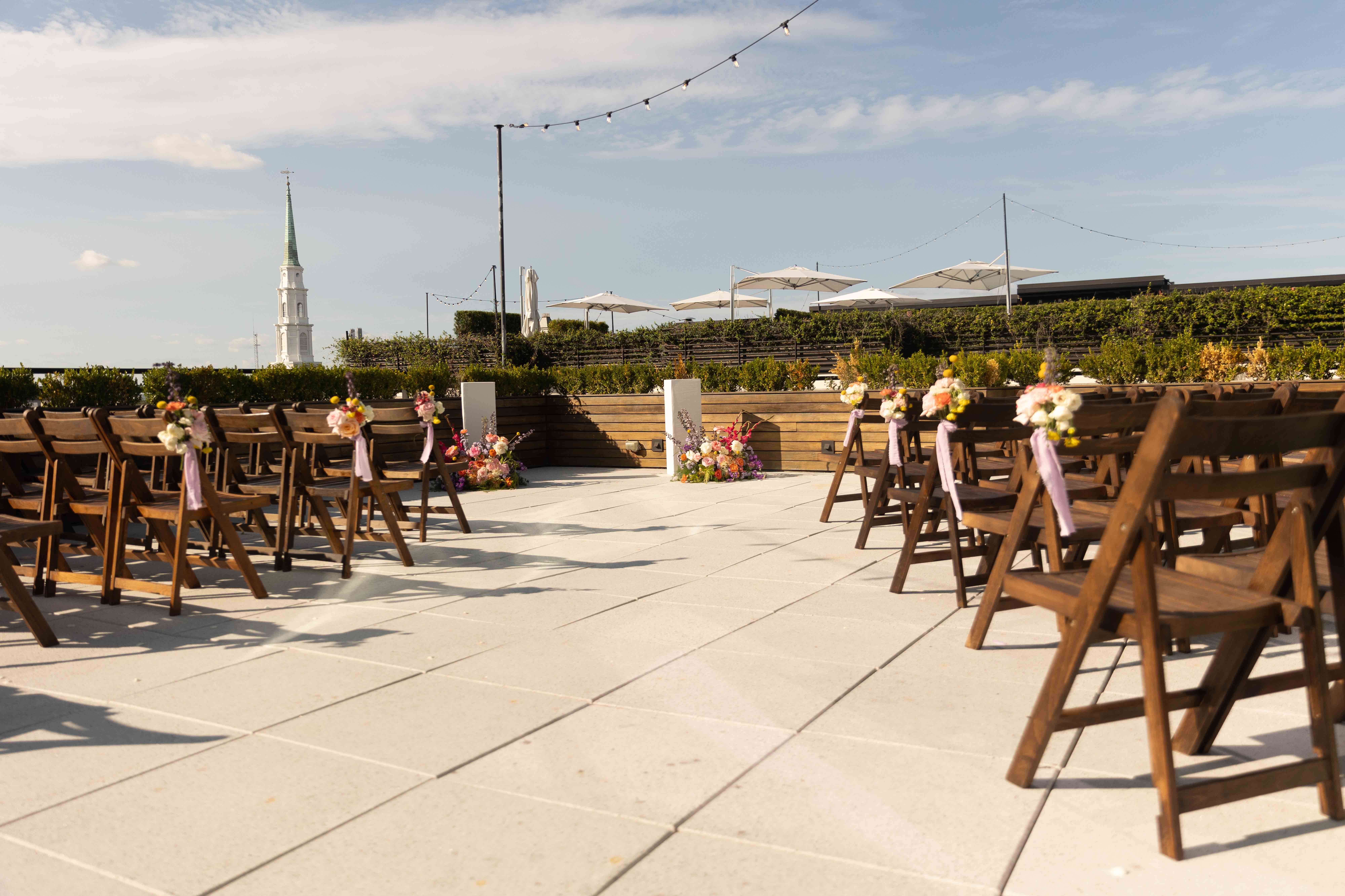 Outdoor wedding ceremony setup with rows of wooden chairs facing an altar adorned with flowers. A white spire is visible in the background under a clear sky at the perry lane hotel