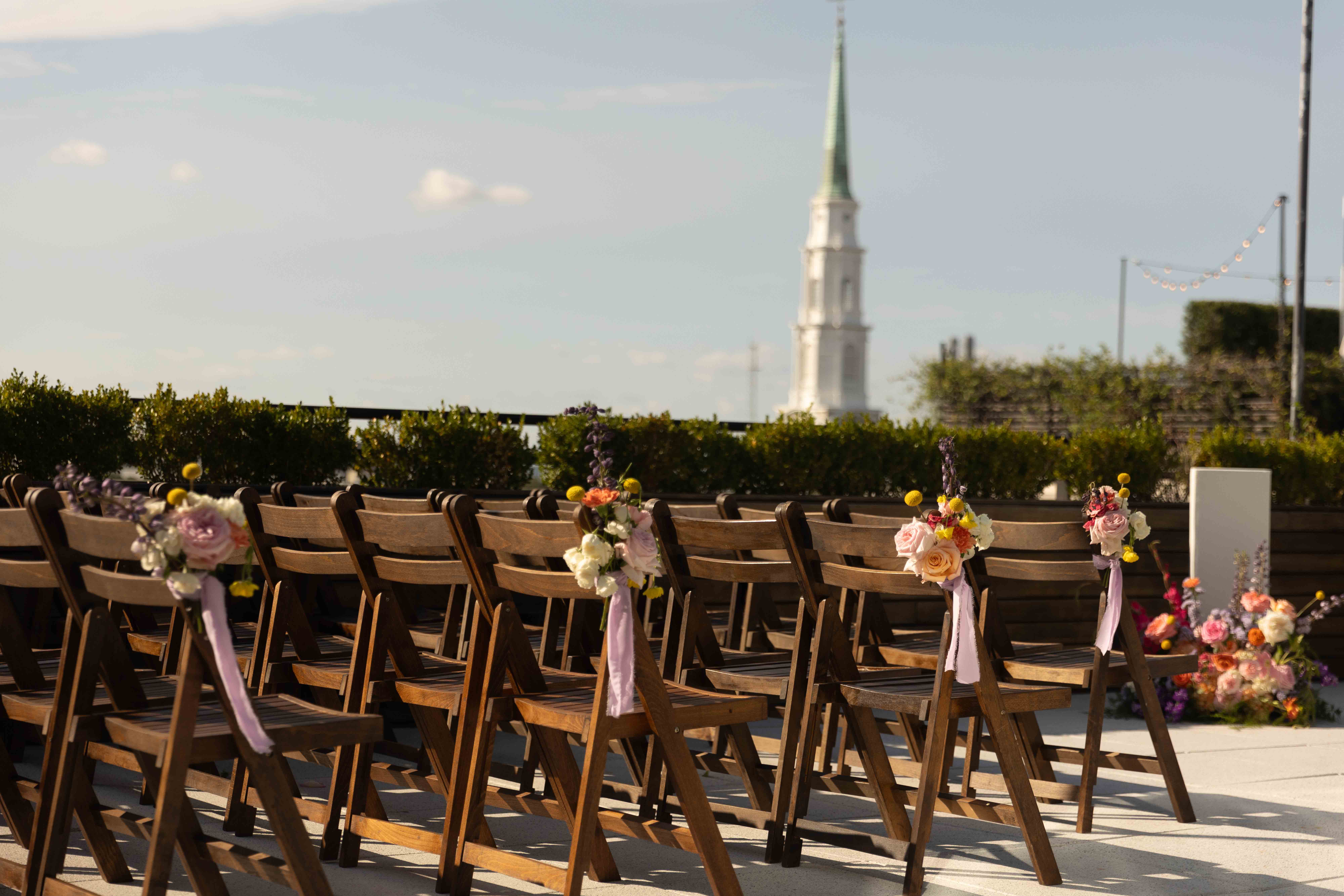 A banquet hall is set up with long tables covered in white tablecloths, decorated with floral centerpieces, and surrounded by white upholstered chairs. Large windows are visible in the background at the perry lane hotel