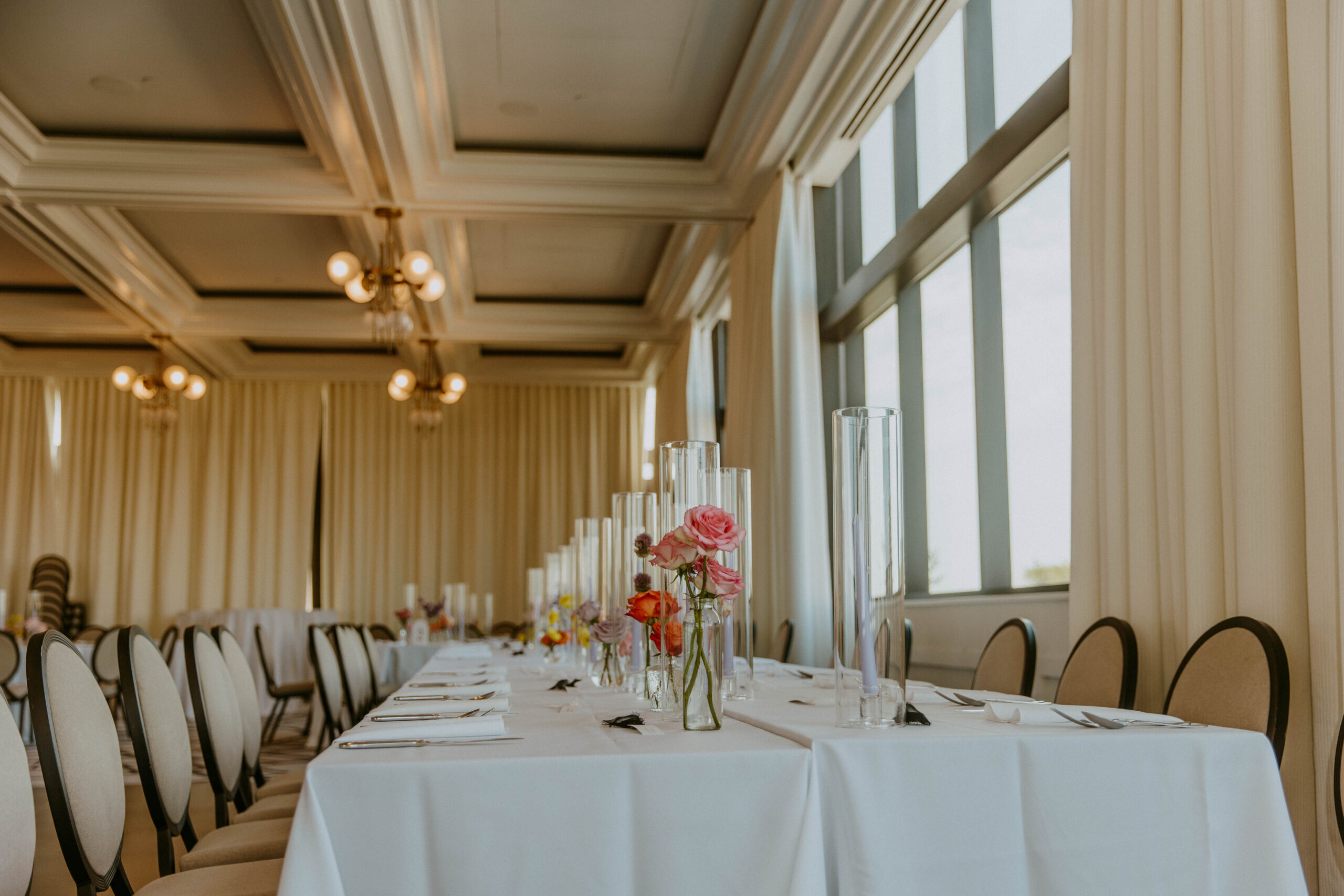 A banquet hall is set up with long tables covered in white tablecloths, decorated with floral centerpieces, and surrounded by white upholstered chairs. Large windows are visible in the background.