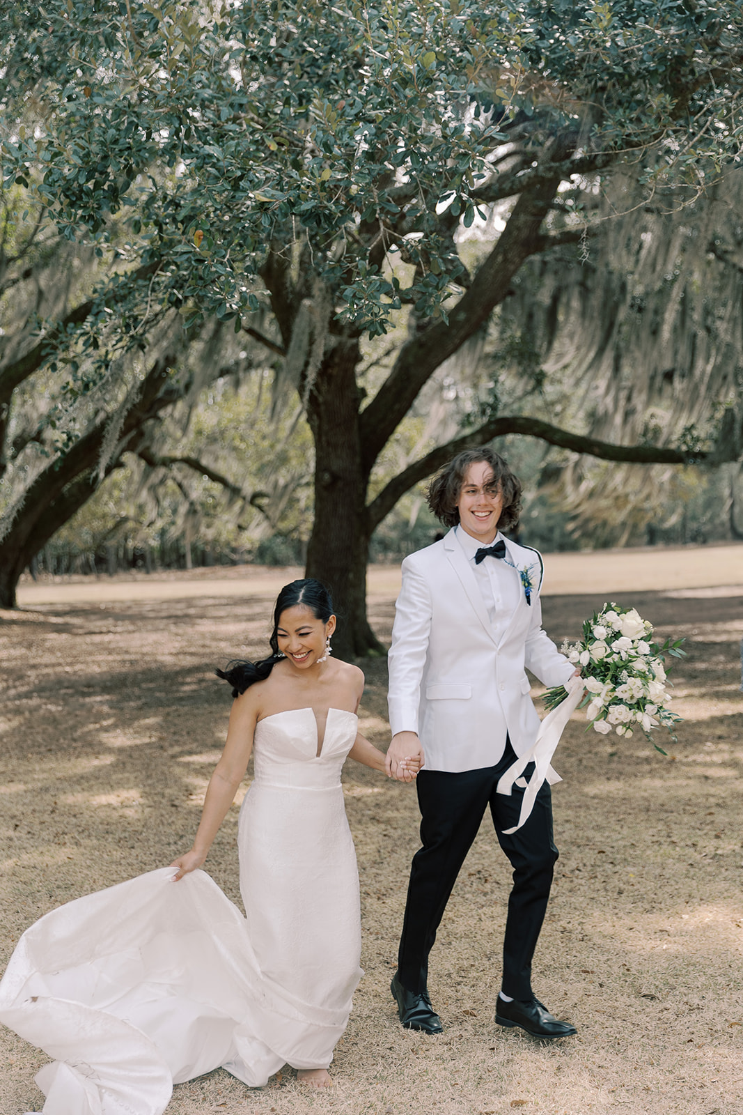 A bride in a strapless white gown and a groom in a white tuxedo walk hand in hand outdoors beneath large trees, with the bride holding her dress and the groom holding a bouquet at their wedding venue Hewitt Oaks