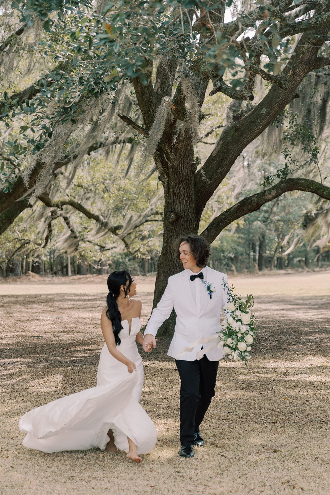 A bride in a strapless white gown and a groom in a white tuxedo walk hand in hand outdoors beneath large trees, with the bride holding her dress and the groom holding a bouquet at their wedding venue Hewitt Oaks