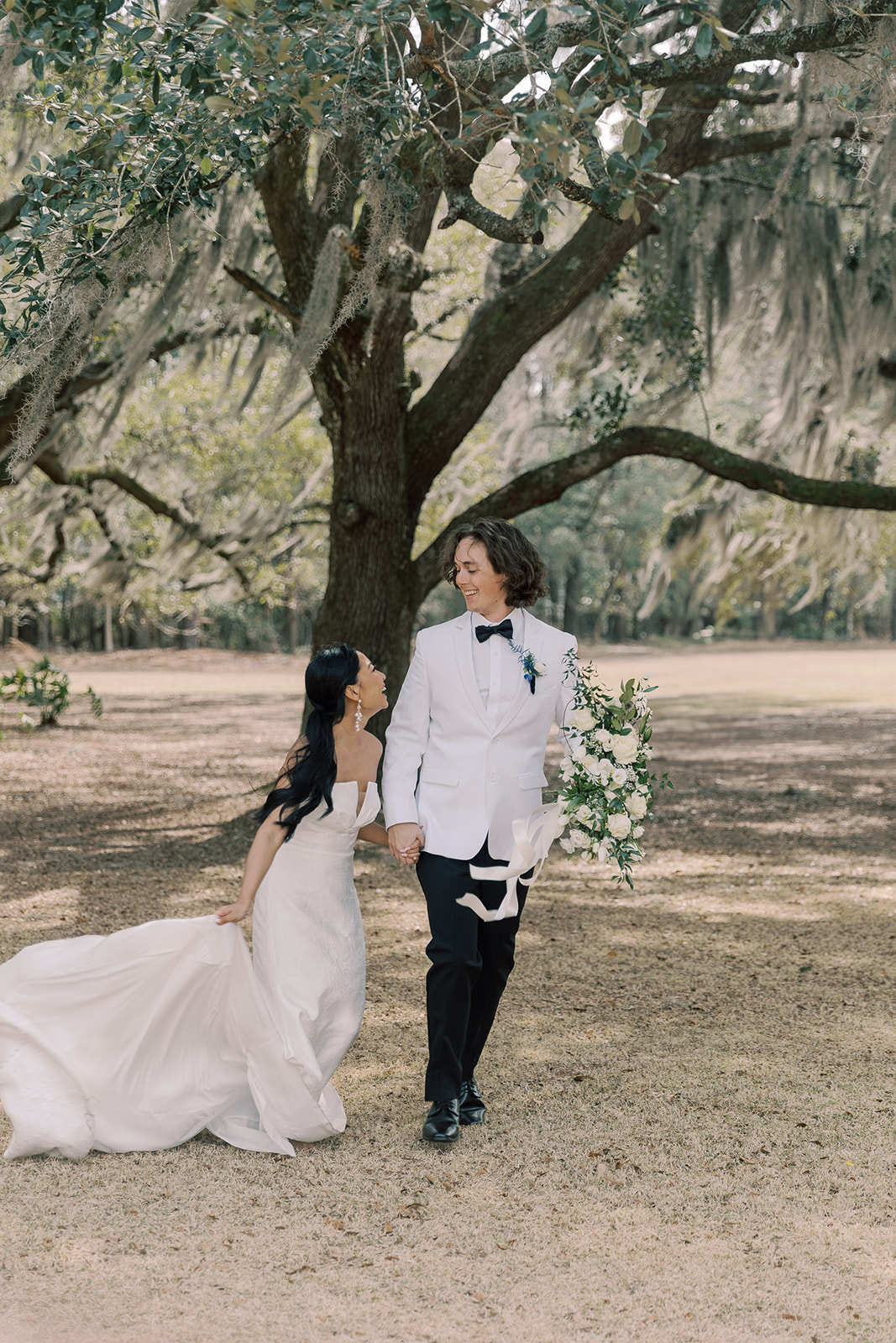 A bride in a strapless white gown and a groom in a white tuxedo walk hand in hand outdoors beneath large trees, with the bride holding her dress and the groom holding a bouquet at their wedding venue Hewitt Oaks