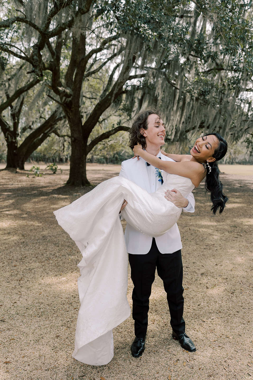 A bride in a strapless white gown and a groom in a white tuxedo walk hand in hand outdoors beneath large trees, with the bride holding her dress and the groom holding a bouquet at their wedding venue Hewitt Oaks