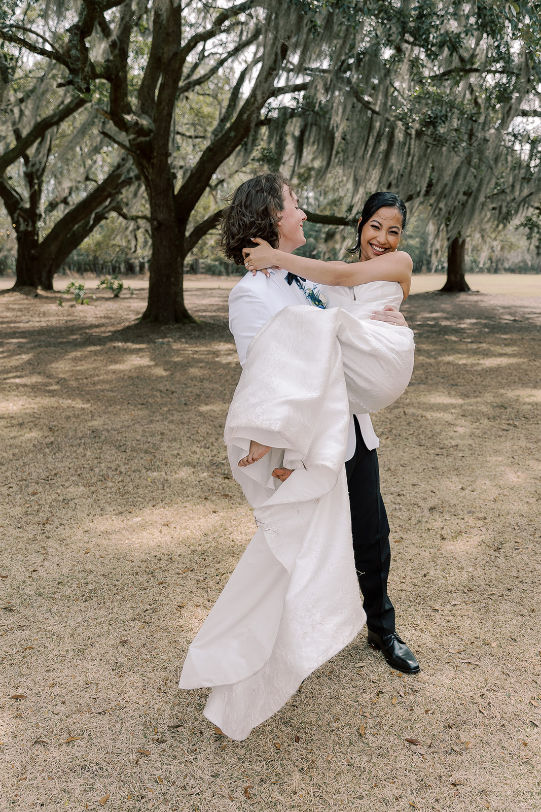 A bride in a strapless white gown and a groom in a white tuxedo walk hand in hand outdoors beneath large trees, with the bride holding her dress and the groom holding a bouquet at their wedding venue Hewitt Oaks