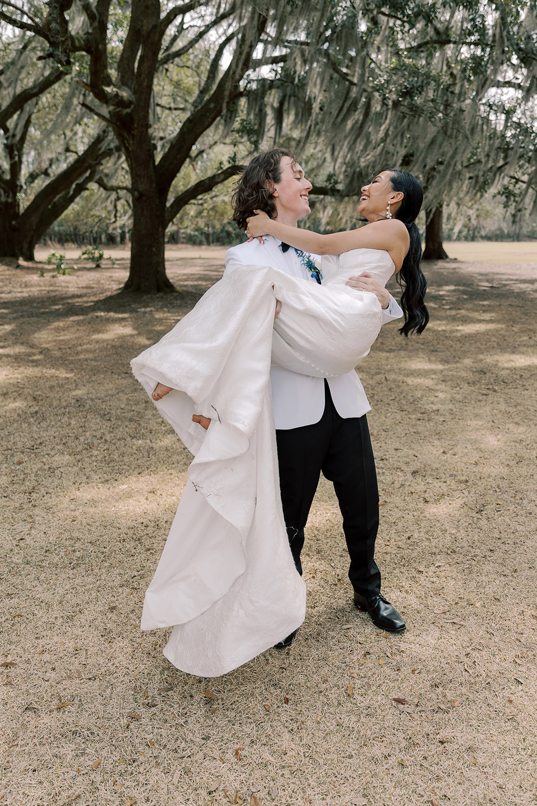 A groom in a white tuxedo lifts and carries a bride in a white wedding dress as they laugh together outdoors under large trees at Hewitt Oaks