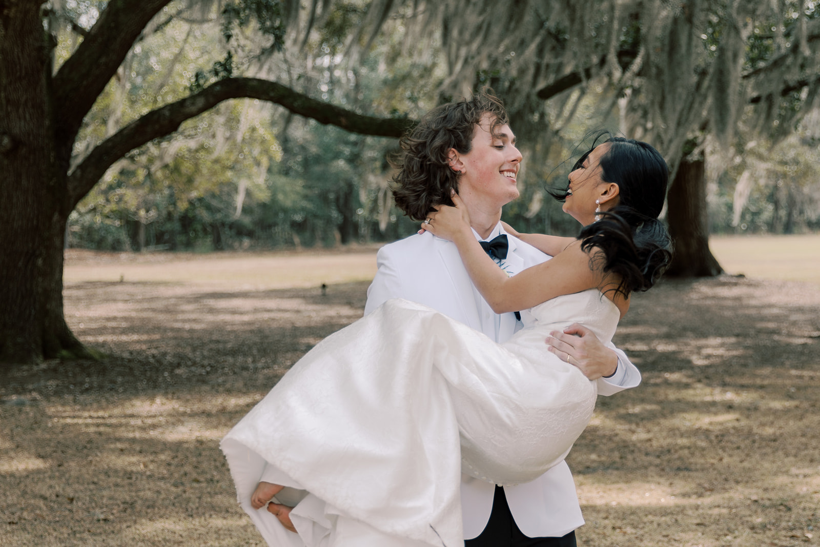 A groom in a white tuxedo lifts and carries a bride in a white wedding dress as they laugh together outdoors under large trees at Hewitt Oaks