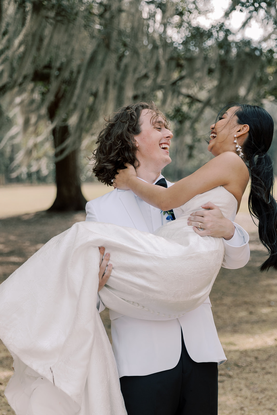 A groom in a white tuxedo lifts and carries a bride in a white wedding dress as they laugh together outdoors under large trees at Hewitt Oaks