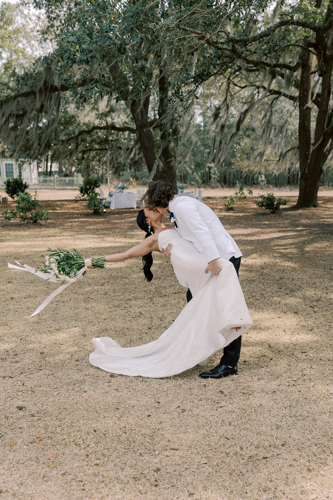 A groom in a white jacket and black pants kisses a bride in a white gown as he dips her backward in a grassy outdoor setting with trees in the background.