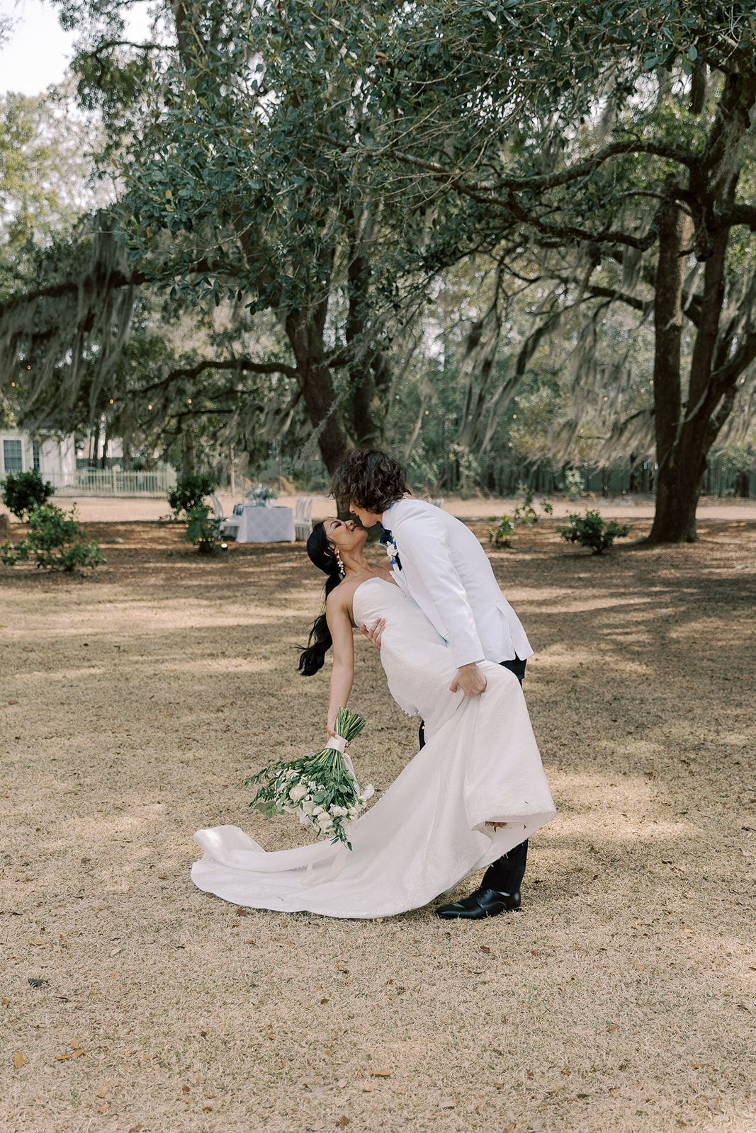 A groom in a white jacket and black pants kisses a bride in a white gown as he dips her backward in a grassy outdoor setting with trees in the background.