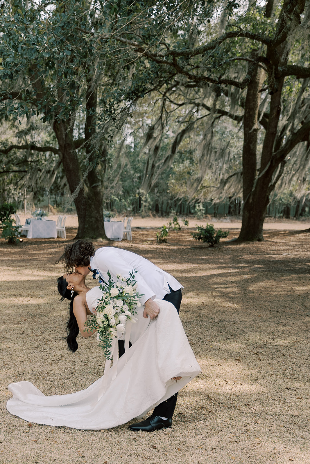 A groom in a white jacket and black pants kisses a bride in a white gown as he dips her backward in a grassy outdoor setting with trees in the background.