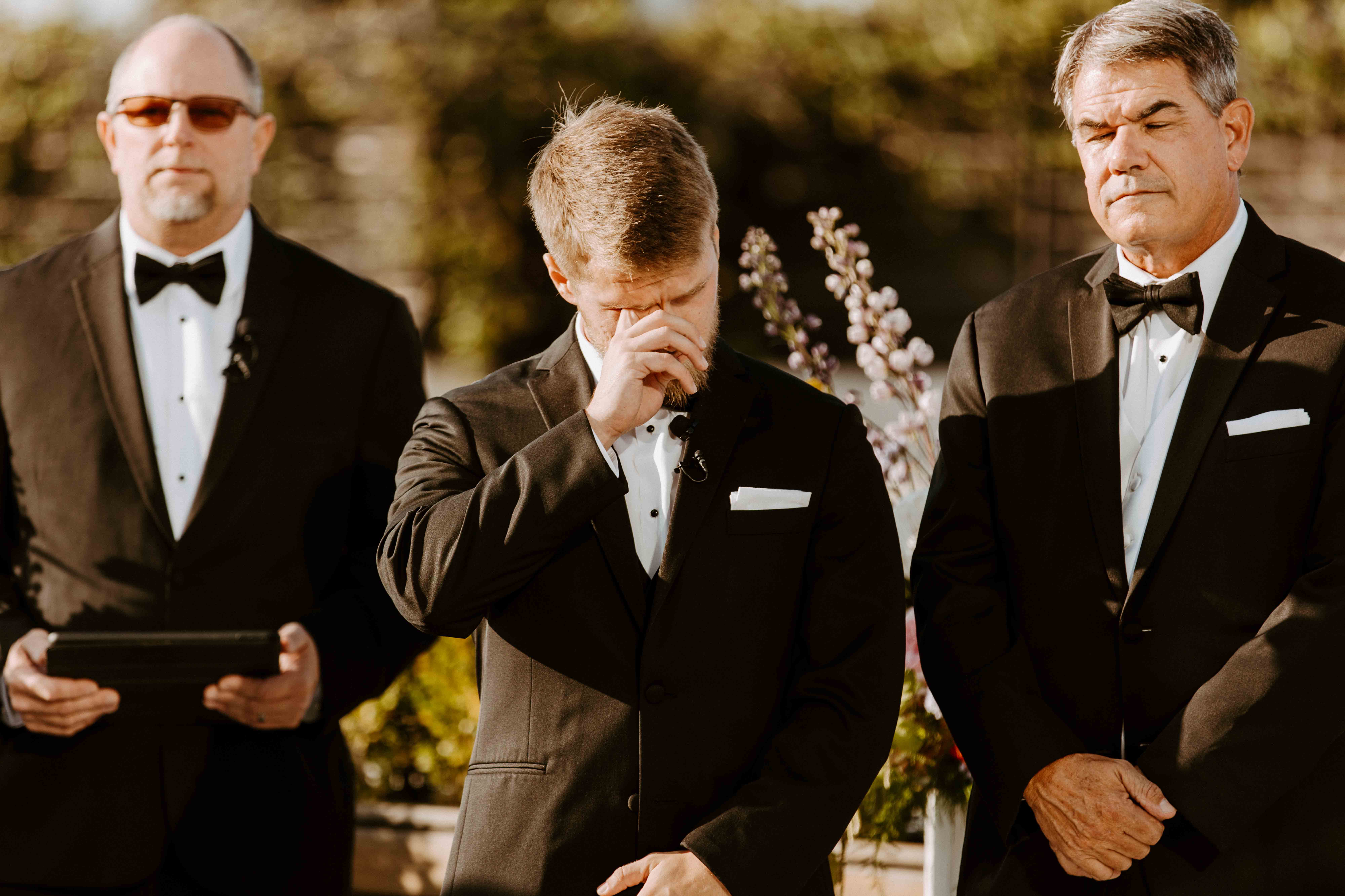 Three men in tuxedos stand outside. The man in the center appears emotional, with his hand covering his eyes. The two men on either side stand calmly at the perry lane hotel