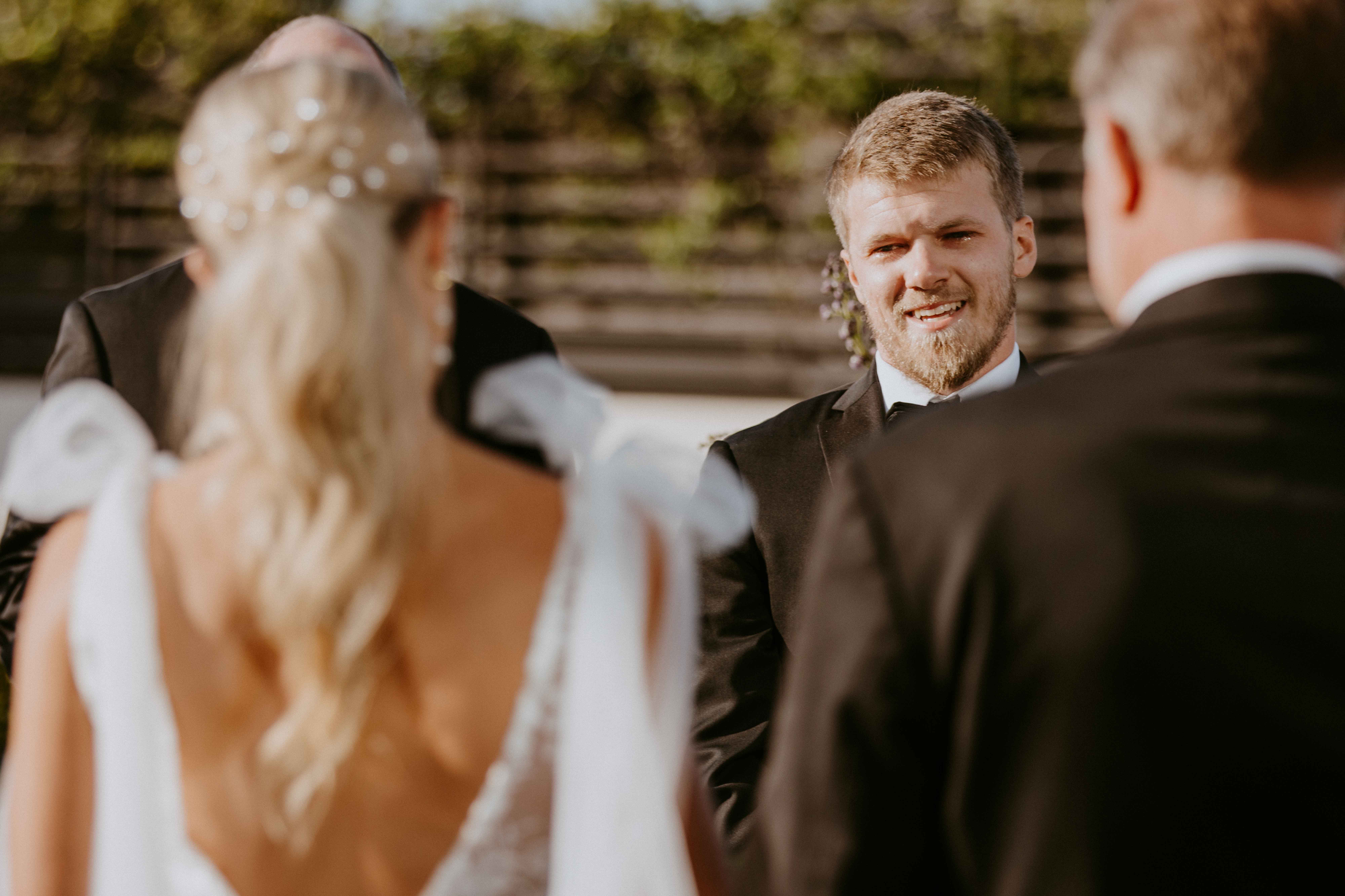 A bride and groom stand facing each other during their wedding ceremony. A man in a suit officiates the ceremony. A colorful floral arrangement decorates the setting at the perry lane hotel