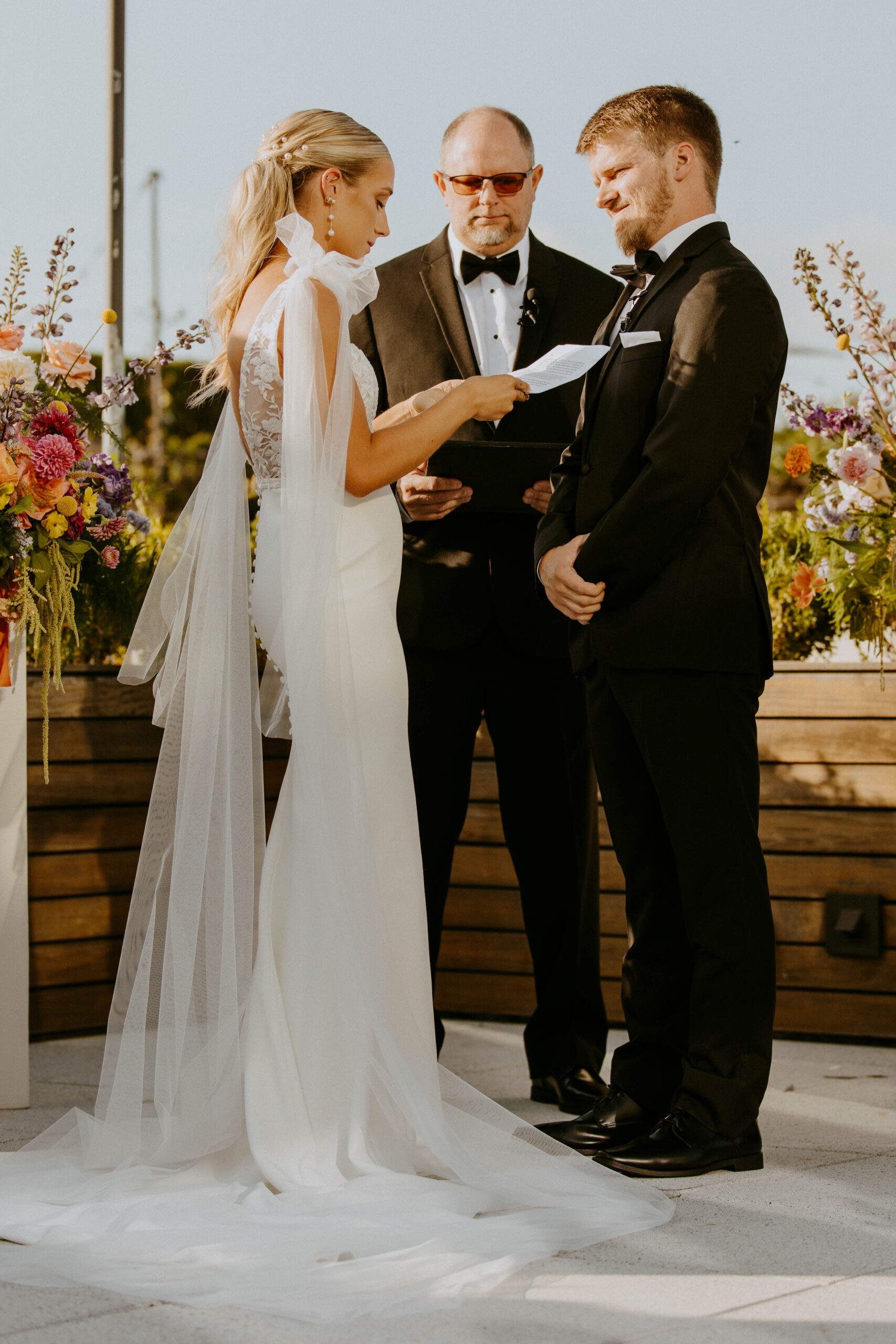 A bride and groom stand facing each other during their wedding ceremony. A man in a suit officiates the ceremony. A colorful floral arrangement decorates the setting at the perry lane hote