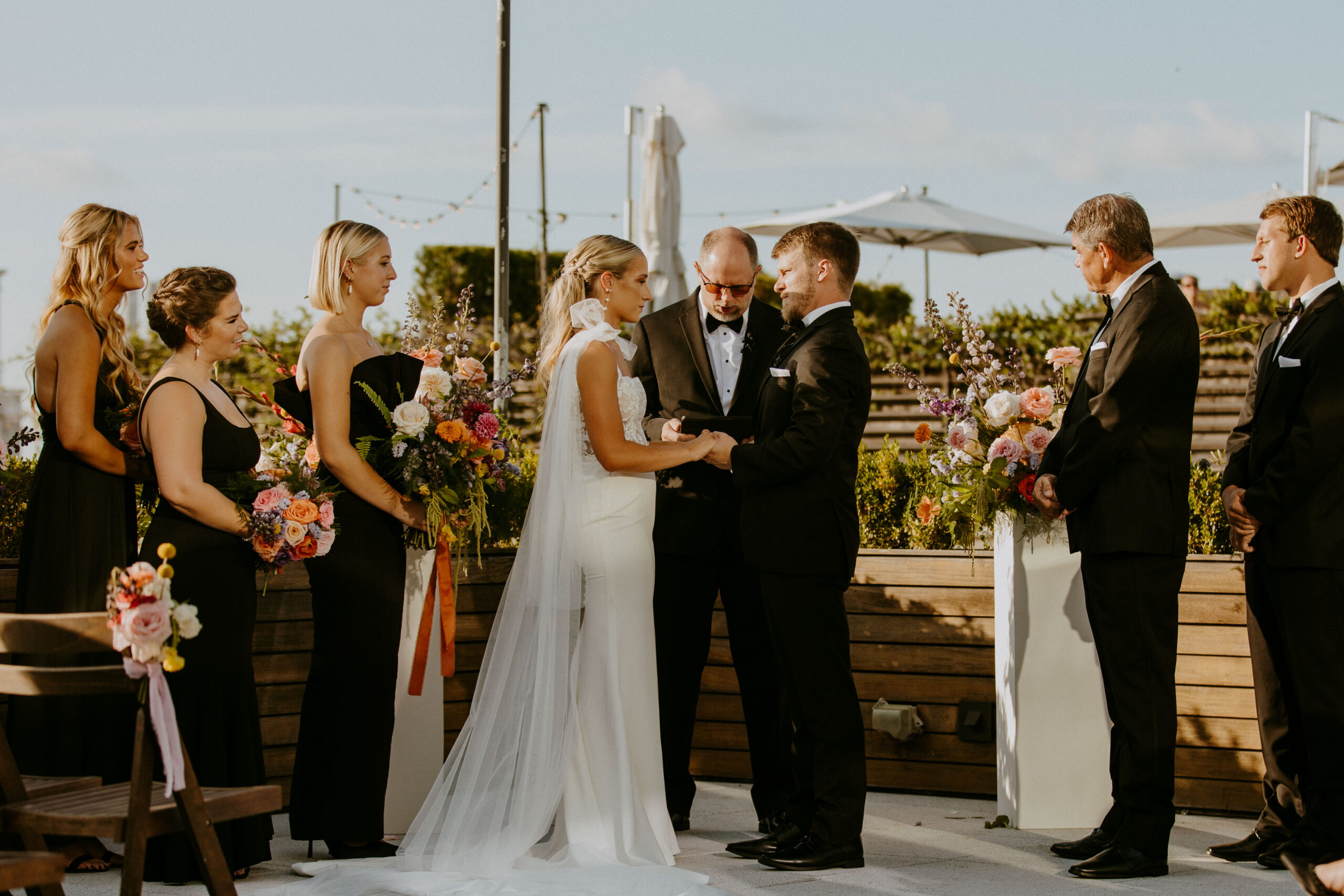 A bride and groom stand facing each other during their wedding ceremony. A man in a suit officiates the ceremony. A colorful floral arrangement decorates the setting at the perry lane hote