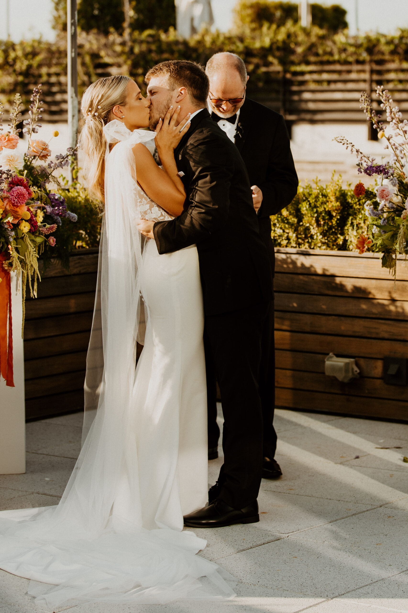 A bride and groom stand facing each other during their wedding ceremony. A man in a suit officiates the ceremony. A colorful floral arrangement decorates the setting at the perry lane hotel