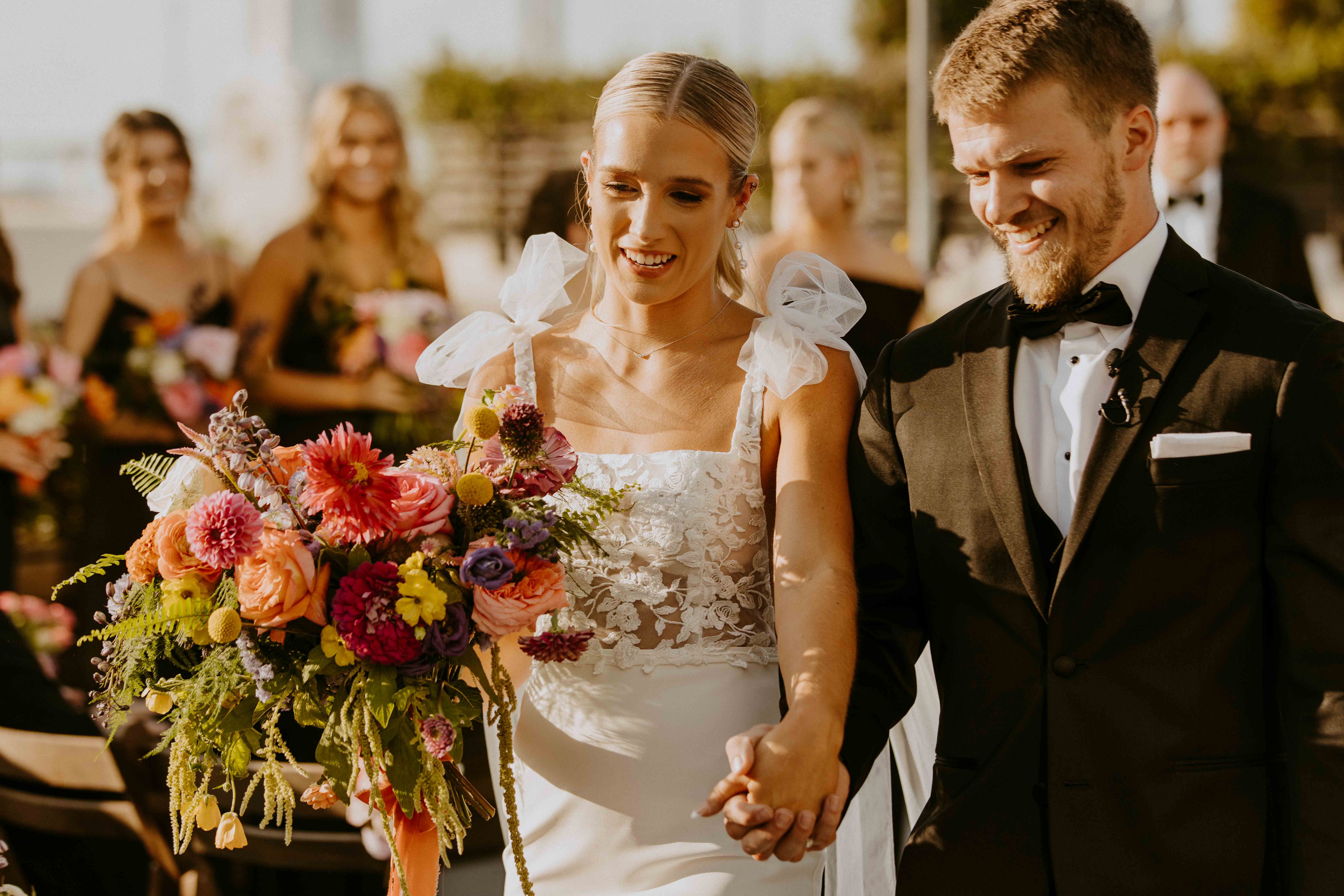 A bride and groom stand facing each other during their wedding ceremony. A man in a suit officiates the ceremony. A colorful floral arrangement decorates the setting at the perry lane hote