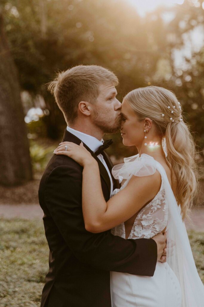 A bride and groom pose outdoors in front of a building with a sign that reads "Savannah." The groom wears a black tuxedo, and the bride wears a white wedding dress, holding a colorful bouquet for their wedding at the the perry lane hotel