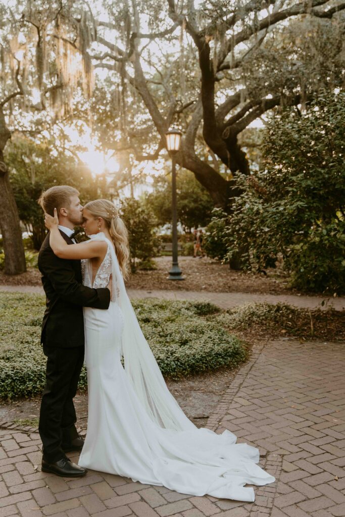 A bride and groom pose outdoors in front of a building with a sign that reads "Savannah." The groom wears a black tuxedo, and the bride wears a white wedding dress, holding a colorful bouquet for their wedding at the the perry lane hotel