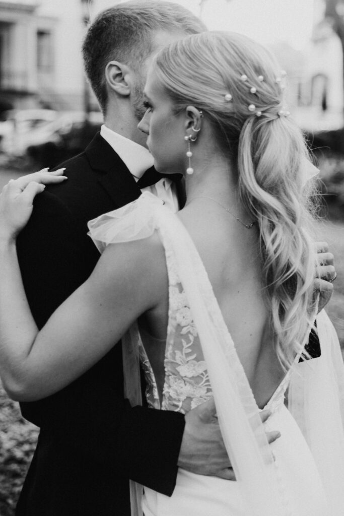 A bride and groom pose outdoors in front of a building with a sign that reads "Savannah." The groom wears a black tuxedo, and the bride wears a white wedding dress, holding a colorful bouquet for their wedding at the the perry lane hotel