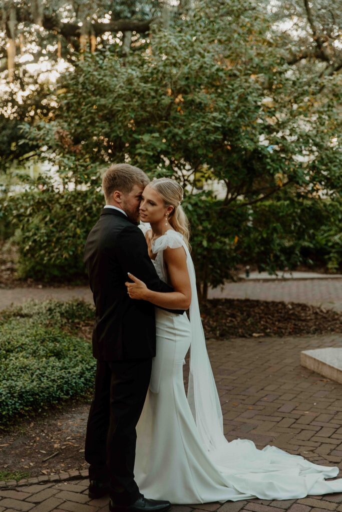 A bride and groom pose outdoors in front of a building with a sign that reads "Savannah." The groom wears a black tuxedo, and the bride wears a white wedding dress, holding a colorful bouquet for their wedding at the the perry lane hotel