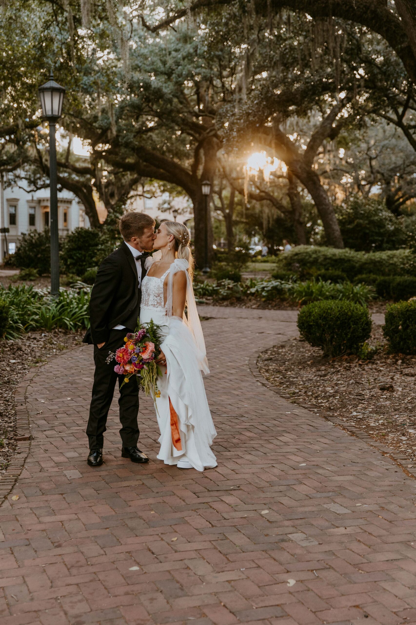 A bride and groom pose outdoors in front of a building with a sign that reads "Savannah." The groom wears a black tuxedo, and the bride wears a white wedding dress, holding a colorful bouquet for their wedding at the the perry lane hotel