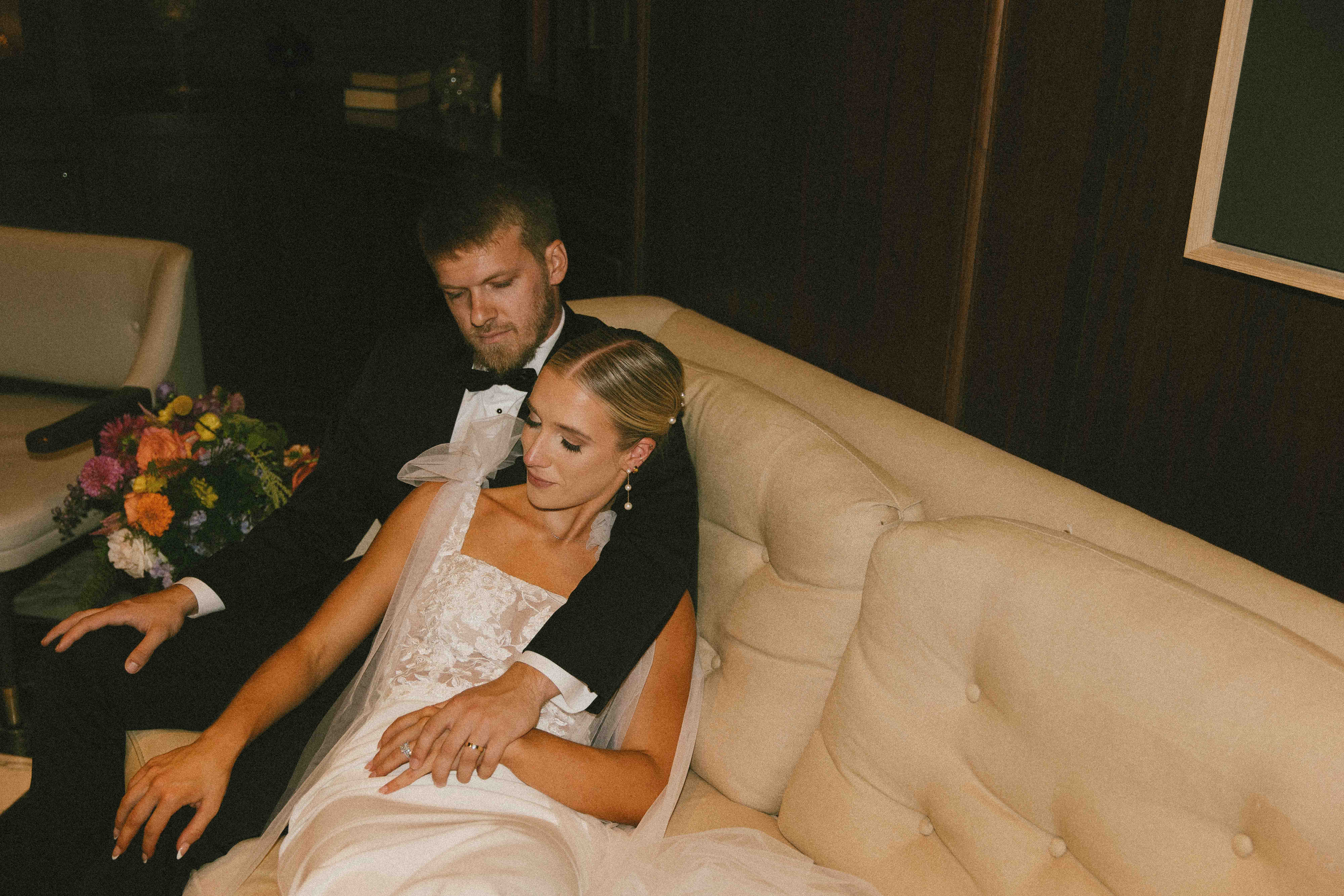 A couple dressed in formal attire, the man in a suit and the woman in a gown, sit on a plush sofa in a room with a framed painting on the wall behind them.