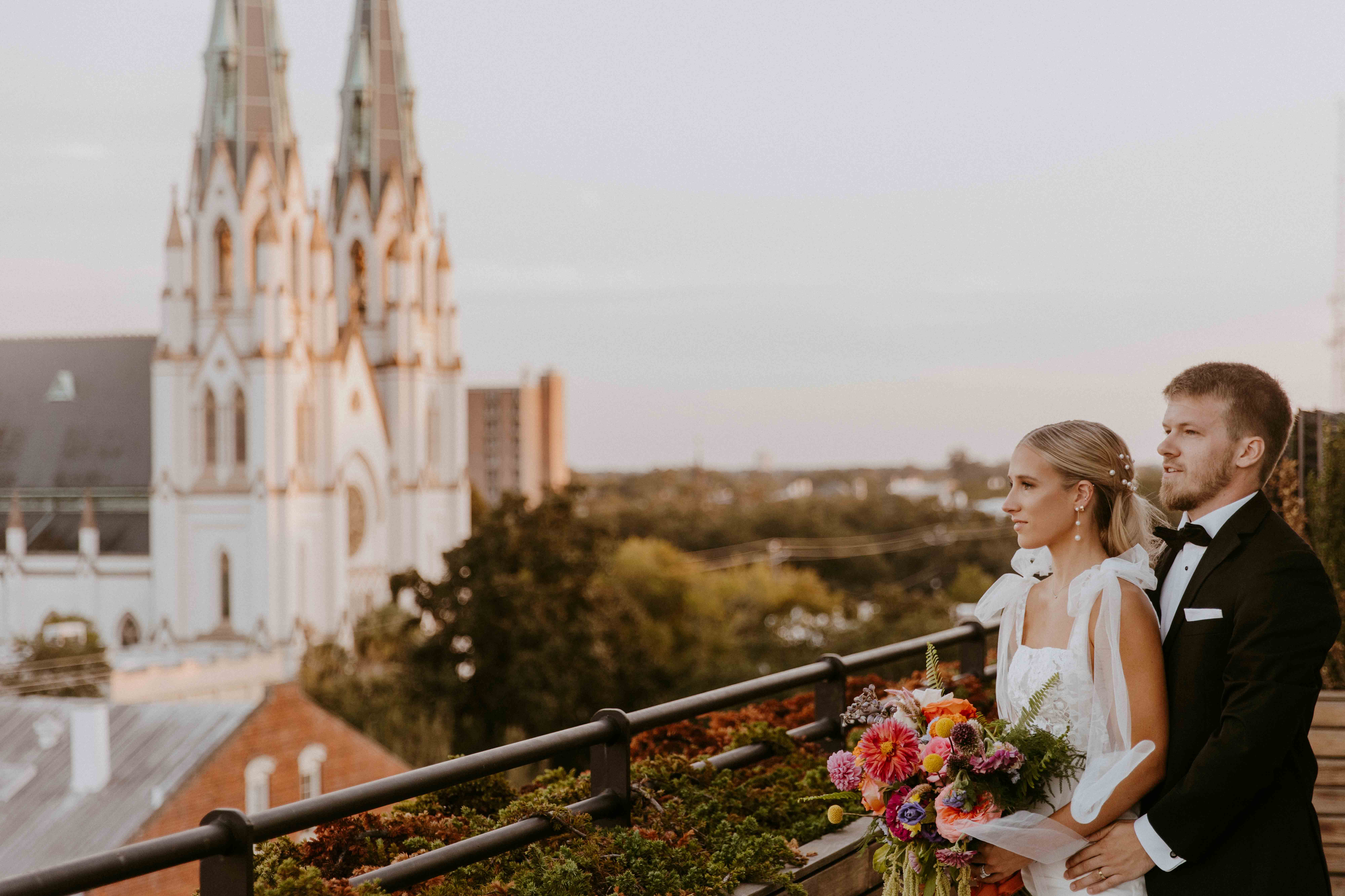 A bride and groom stand on a balcony overlooking a landscape with a large church in the background. The bride holds a vibrant bouquet at the perry lane hotel