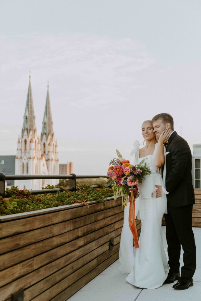 A bride and groom in wedding attire stand on a rooftop adorned with flowers and plants, with a pair of church spires visible in the background. The groom gently touches the bride's face.