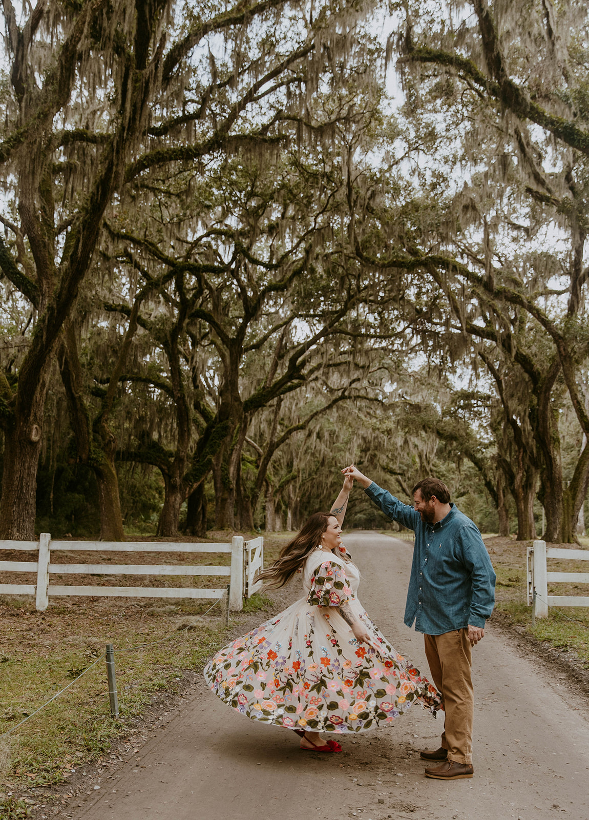 A couple dances on a tree-lined dirt path, with the woman wearing a flowing floral dress and the man in casual attire, surrounded by rustic wooden fences 