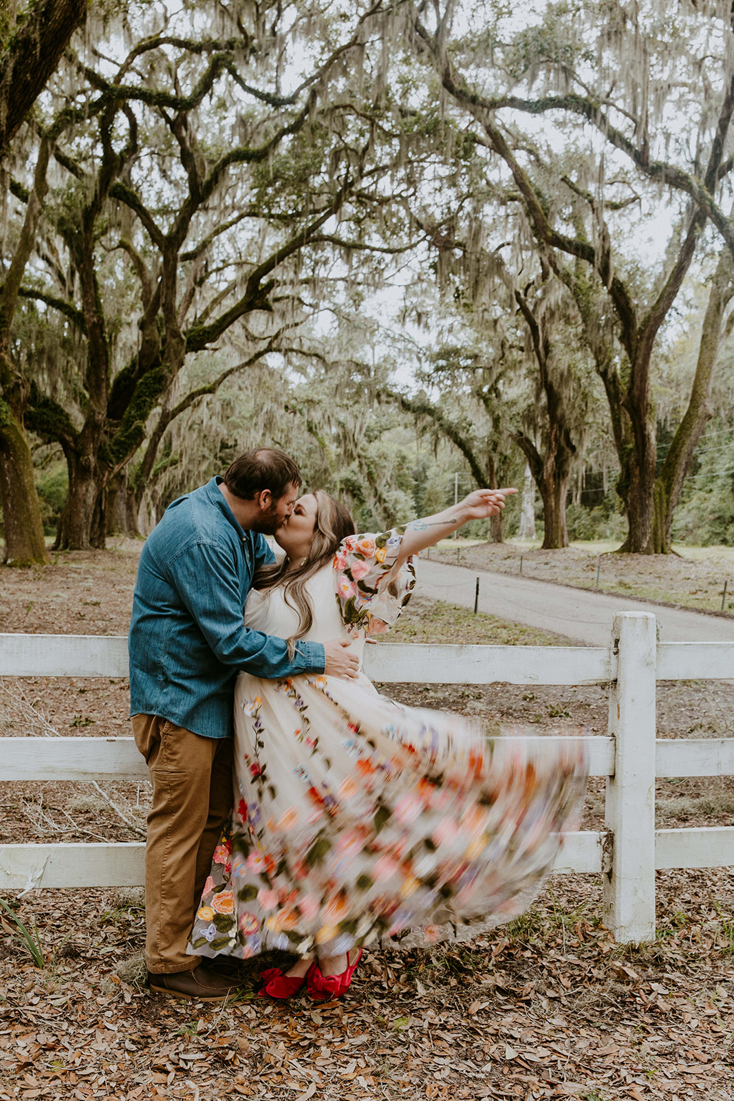 A couple dances on a tree-lined dirt path, with the woman wearing a flowing floral dress and the man in casual attire, surrounded by rustic wooden fences for their savannah engagement session