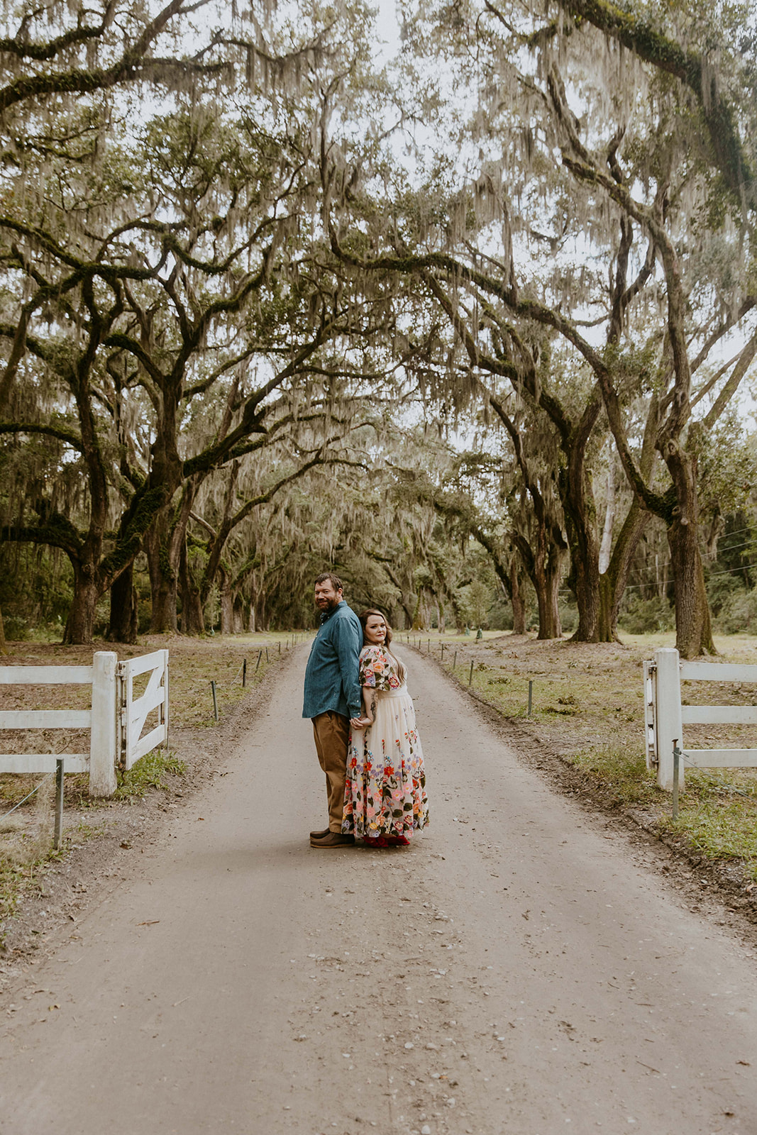 A couple dances on a tree-lined dirt path, with the woman wearing a flowing floral dress and the man in casual attire, surrounded by rustic wooden fences for their savannah engagement session